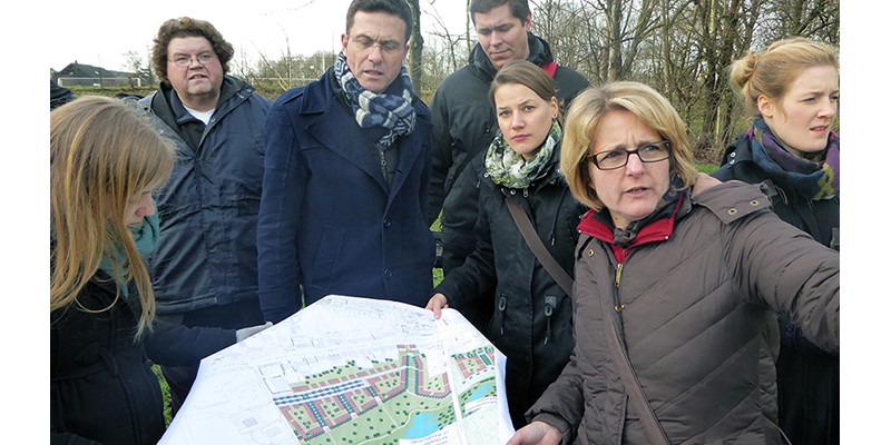 Das Projektteam hält gemeinschaftliche eine Karte des zukünftigenOstparks in den Händen und schauen abwechseln auf die Karte und in die Richtung, wo sich das Ziel befindet. Im Hintergrund befinden sich unbelaubte Bäume. Die Personen sind warm angezogen.