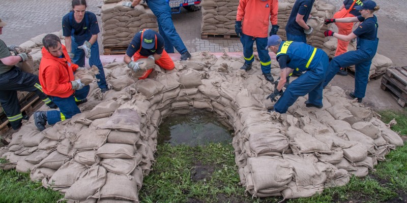 Ausbildungsteilnehmer_innen in Blau gekleidet und mit gelbem Schriftzug THW schichten übungsweise eine Sandsack-Quellkade auf zur Sicherung eines Deiches.