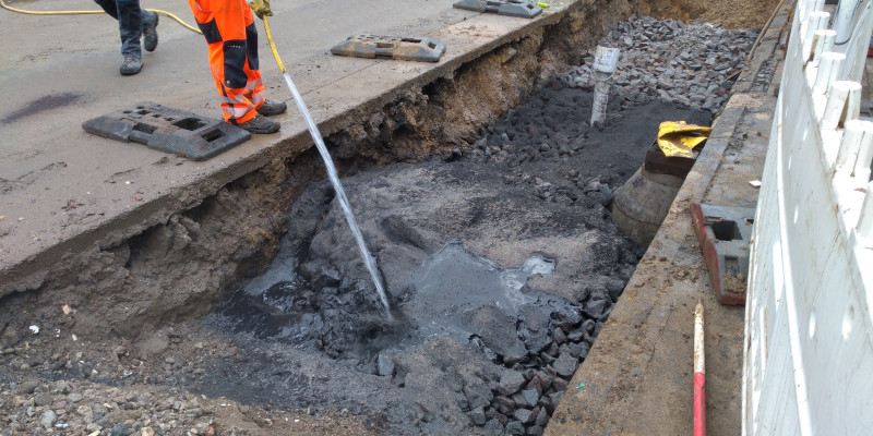 Fotografie einer Baustelle. In einem aufgerissenen Teilabschnitt der Straße wird ein dunkles Gemisch bewässert.