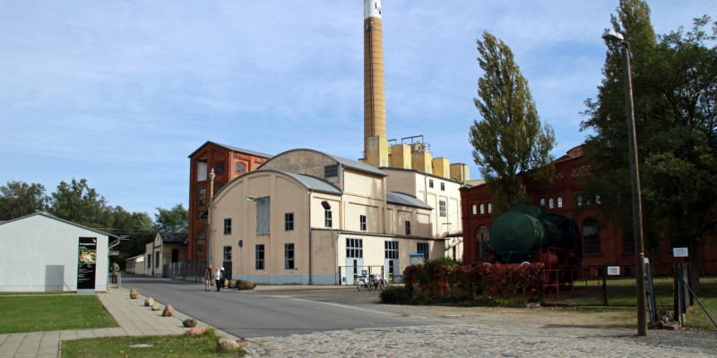 Bildungs- und Transformationszentrum Brikettfabrik LOUISE. Das historische Gebäude hat eine helle Fassade und grau abgesetzte Fenster und runde Dächer. Das Gebäude steht an der geteeerten Fahrbahn an der äußeren Kurve. Es stehen mehrere Gebäude darum herum. Im Hintergrund ist der blaue Himmel sowie ein Schornstein zu sehen.