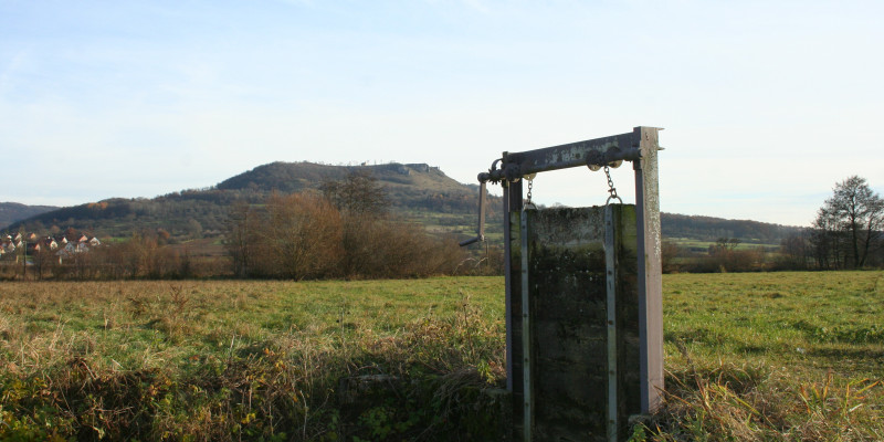 Mittig im Bild steht eine Kurbelschütz (Regulierungsvorrichtung mit Kurbel). Die senkrechte Platte, die den Graben absperren würde, ist hochgekurbelt.
