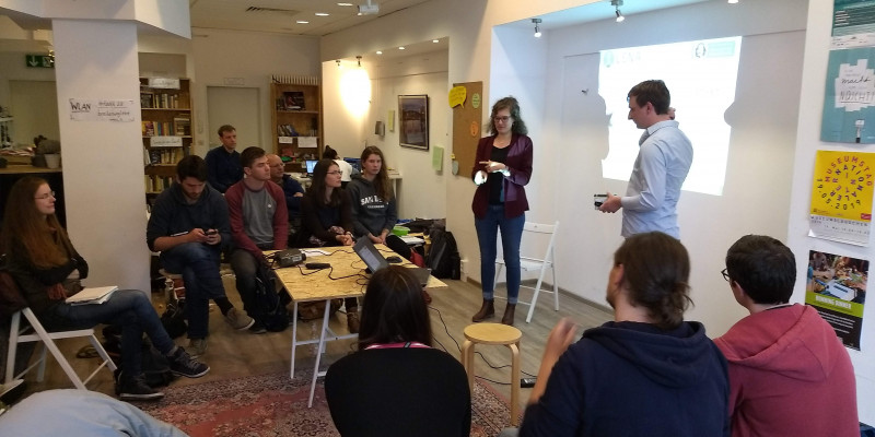 Vor einer Präsentation an der Wand stehen zwei Personen. Sie präsentieren mehreren anderen Personen, die im Halbkreis sitzen etwas.