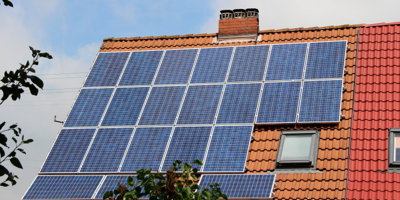 Der Blick geht auf ein rotes Ziegeldach, welches überwiegend mit Solarpaneelen bestückt ist. Im Hintergrund ist bewölkter Himmel zu sehen.