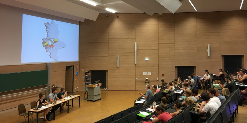 Blick in einen großen Hörsaal. Vorne sitzen drei Personen an einem Tisch, daneben eine Person an einem Rednerpult. Ihnen gegenüber sitzen weitere Personen in den Hörsaalrängen.