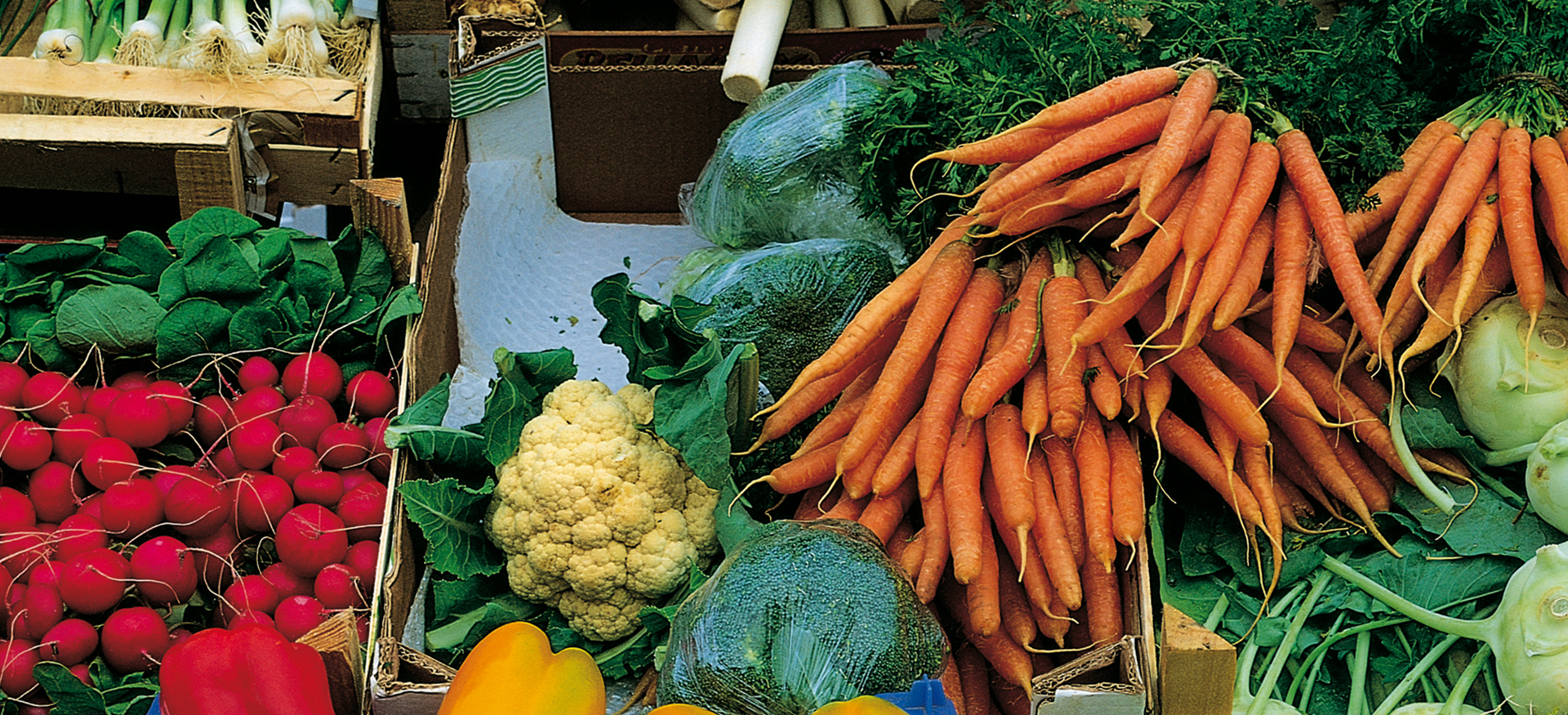 verschiedenes Gemüse (Moehren, Radieschen, Paprika, Zwiebeln, Lauch) werden in Holzkästen auf dem Markt angeboten