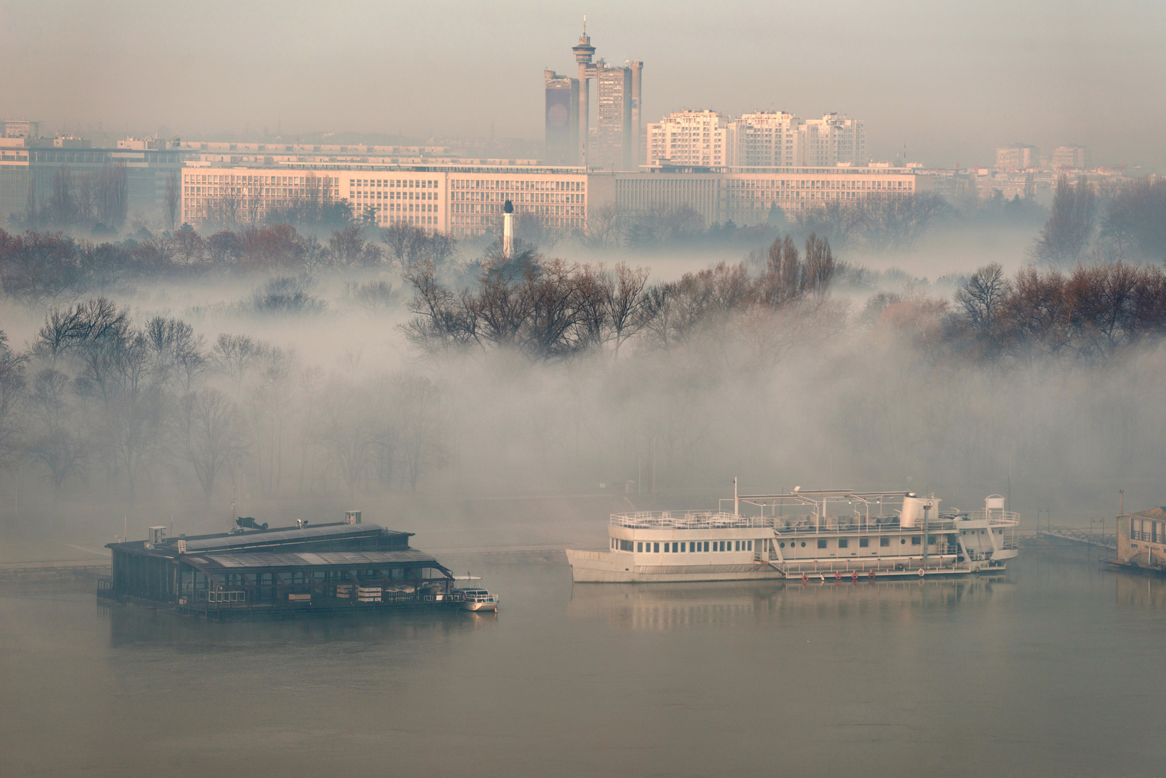 Foto: Nebel über einem Fluss und seinen Uferbereichen. Im Hintergrund sind die Häuser einer Stadt zu sehen.