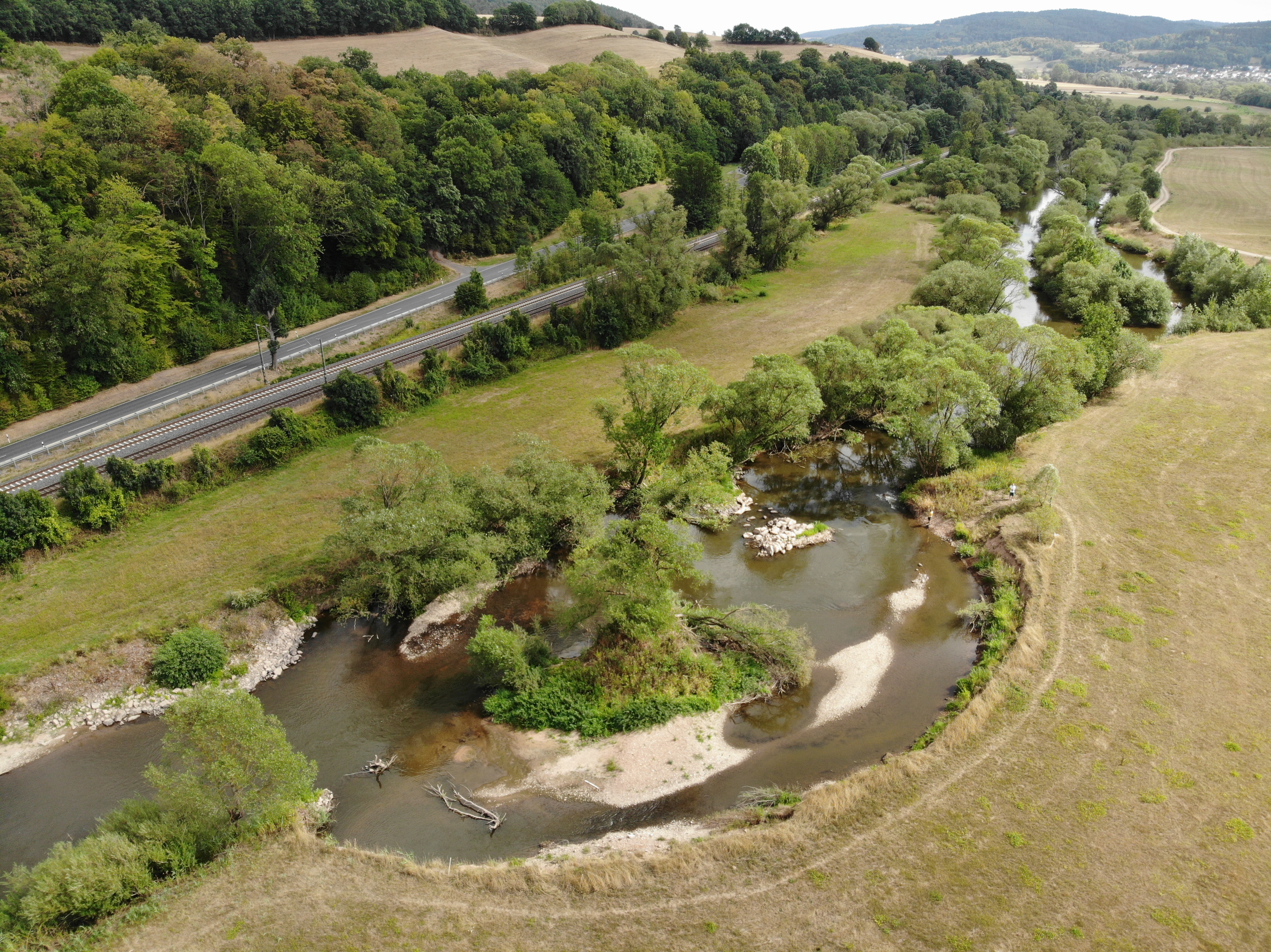 Luftbild der Fulda mit großzügiger Aufweitung und Kiesbänken, die teilweise mit Vegetation bedeckt sind. Links ist eine Straße zu erkennen, rechts grenzt Grünland an das Gewässer an.