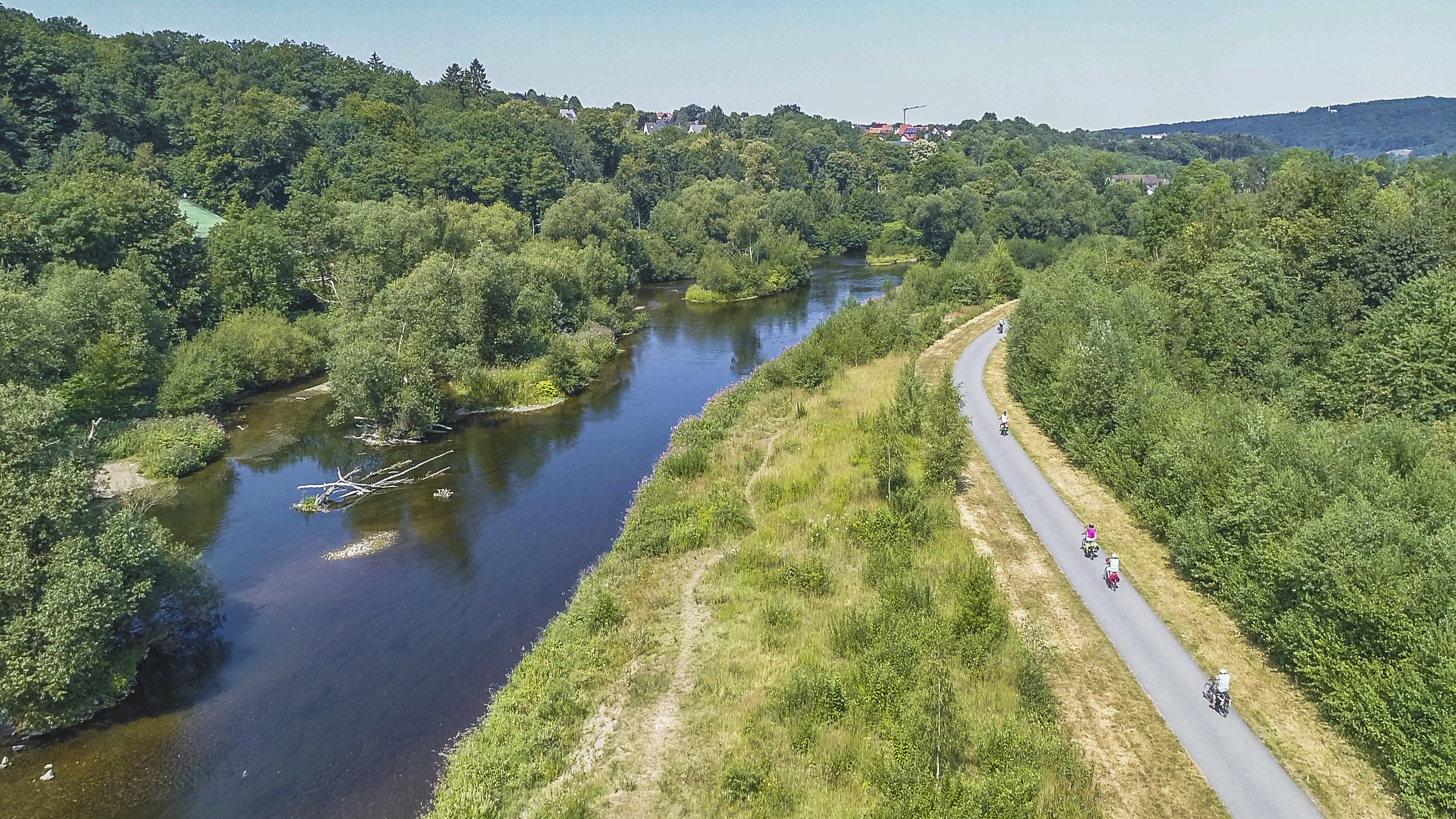 Foto: Der Fluss Ruhr bei Arnsberg mit zahlreichen Inseln, Kiesbänken, Totholz und dichtem Bewuchs. Etwas abseits des renaturierten Flusses verläuft ein befestigter Weg auf dem mehrere Personen auf Rädern unterwegs sind.