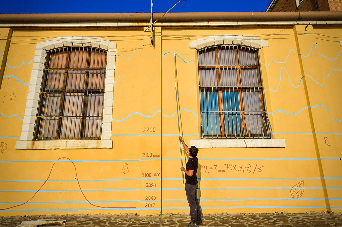 Das Foto zeigt den Künstler Andreco bei der Bemalung einer Hauswand in Venedig. Die Hauswand trägt bereits einige fiktive Hochwassermarken bis 2100. Das Bild visualisiert das Risiko des globalen Meeresspiegelanstiegs für den Menschen jenseits einer globalen Erwärmung auf 1,5 °C. 