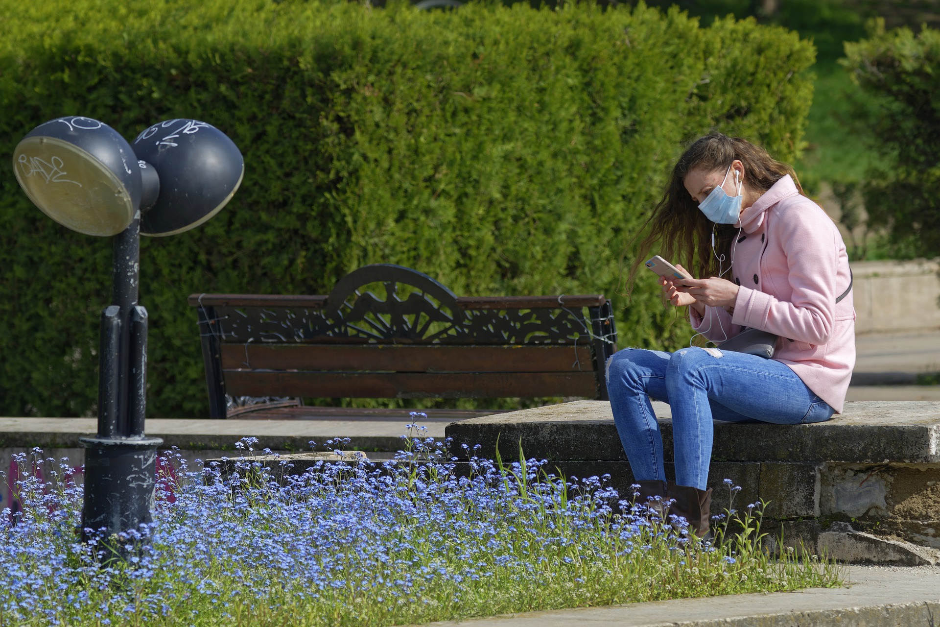 Eine Frau sitzt in einem Park auf einer Bank, trägt einen Mund-Nasen-Schutz und schaut auf ihr Smartphone