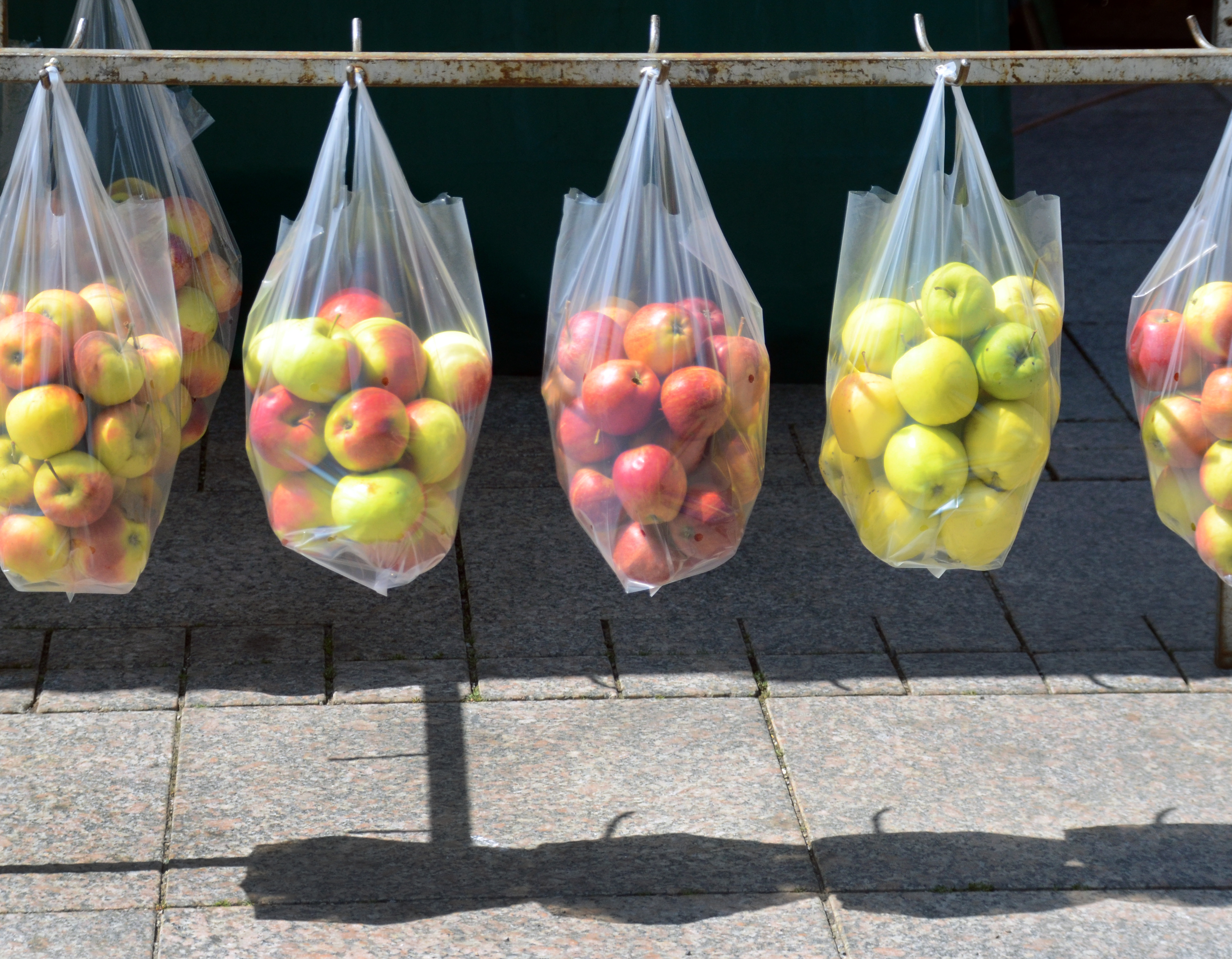 an einer Eisenstange über einem gepflasterten Platz oder Fußweg hängen durchsichtige Plastiktüten mit Äpfeln