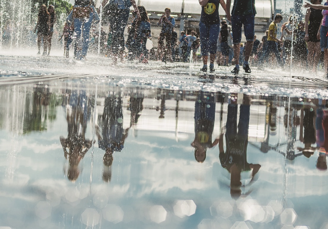 Das Bild zeigt eine Wasserfläche in einer Stadt, offensichtlich ein Brunnen. Es laufen mehrere Menschen über die Wasserfläche und spiegeln sich darin.