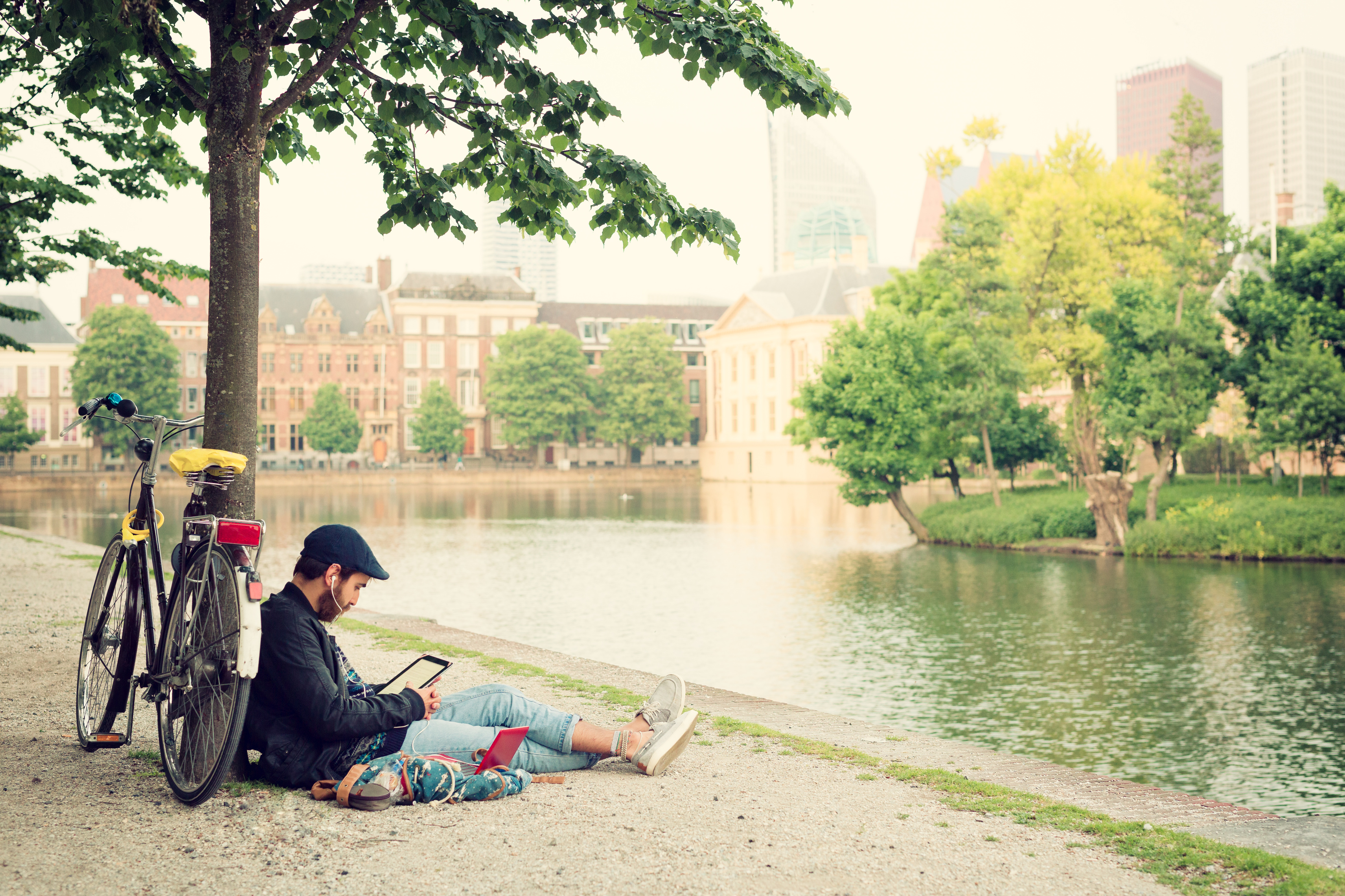 Ein junger Mann sitzt in der Stadt an einem Fluss unter einem Baum und schaut auf sein Tablet mit Kopfhörern im Ohr. Am Baum lehnt sein Fahrrad.