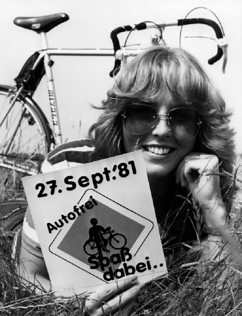eine Frau lmit Sonnenbrille iegt neben ihrem Fahrrad auf einer Wiese und hält ein Schild in die Kamera: 27. Sept.´81, Autofrei – Spaß dabei“