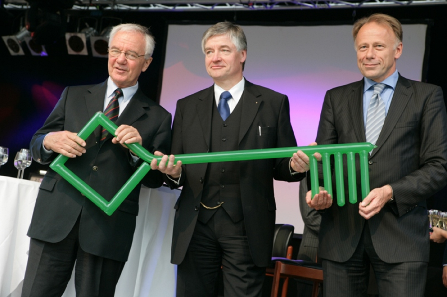 Gruppenfoto: drei Männer in Anzug und Krawatte halten einen riesigen grünen Schlüssel, im Hintergrund Sektgläser