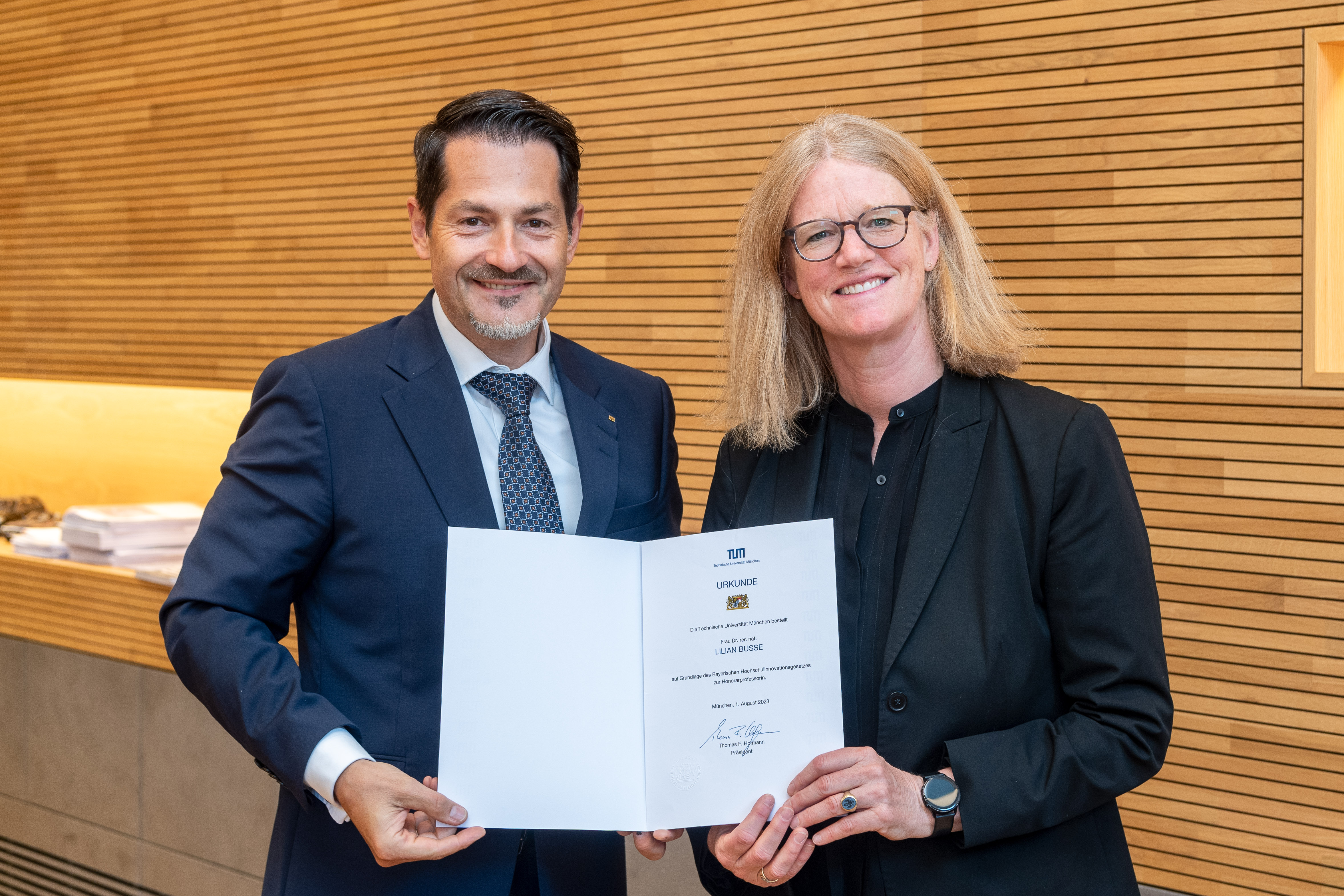 TUM-Präsident Prof. Dr. Thomas Hoffmann und Dr. Lilian Busse halten zusammen eine Urkunde für ein Foto