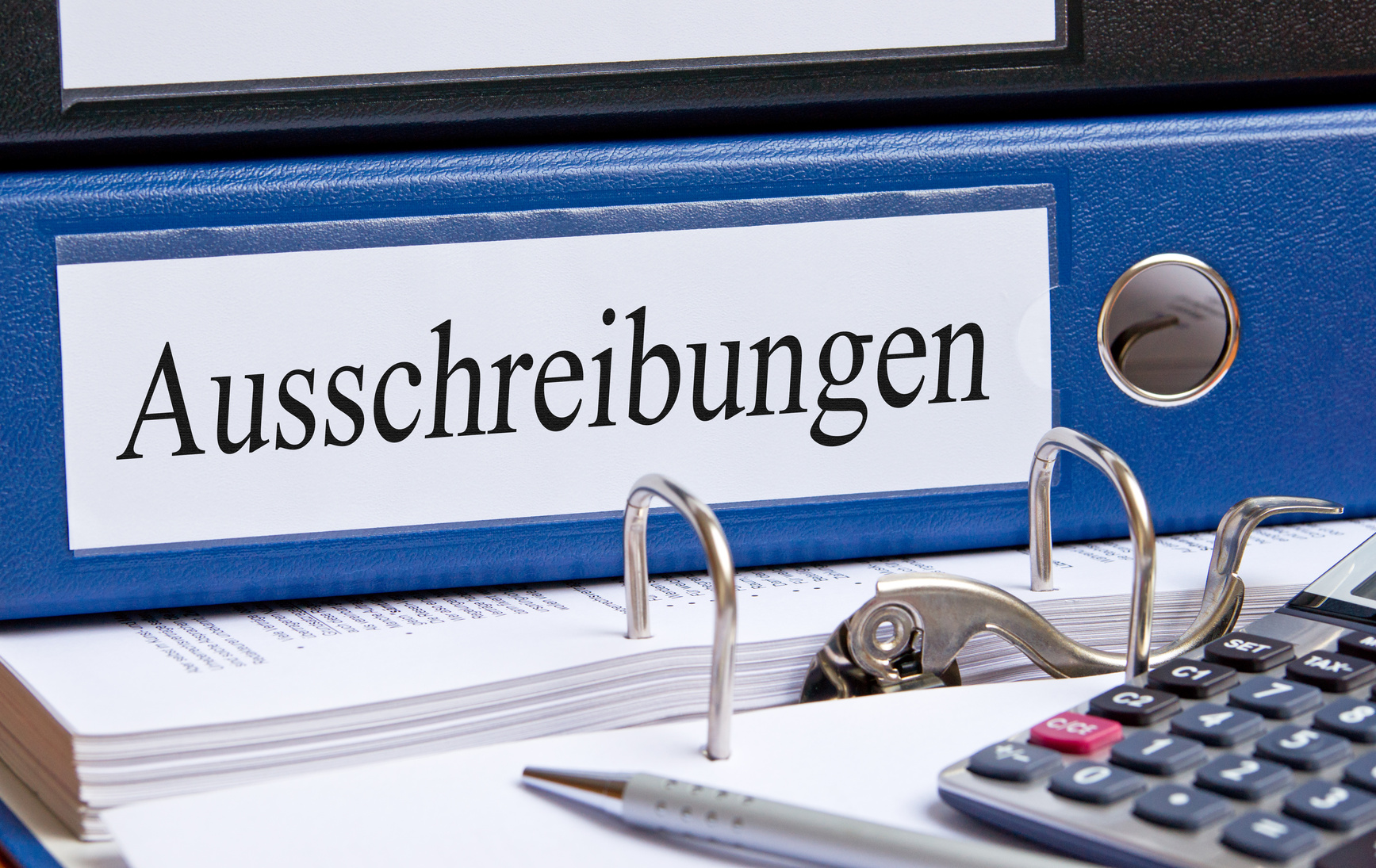 auf einem Tisch liegen ein Ordner mit der Aufschrift "Ausschreibungen", ein Taschenrechner und ein Kugelschreiber