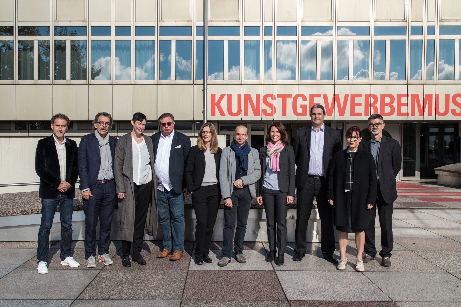 10 Männer und Frauen haben sich vor einem Gebäude mit der Aufschrift "Kunstgewerbemuseum" für ein Gruppenfoto aufgestellt
