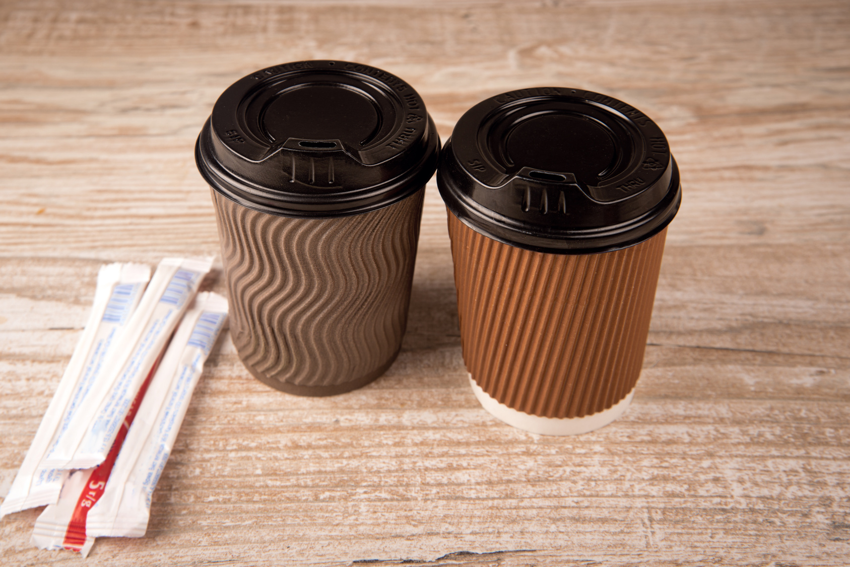 zwei „Coffee to go“-Becher auf einem Tisch, daneben zwei Papiertütchen mit Zucker