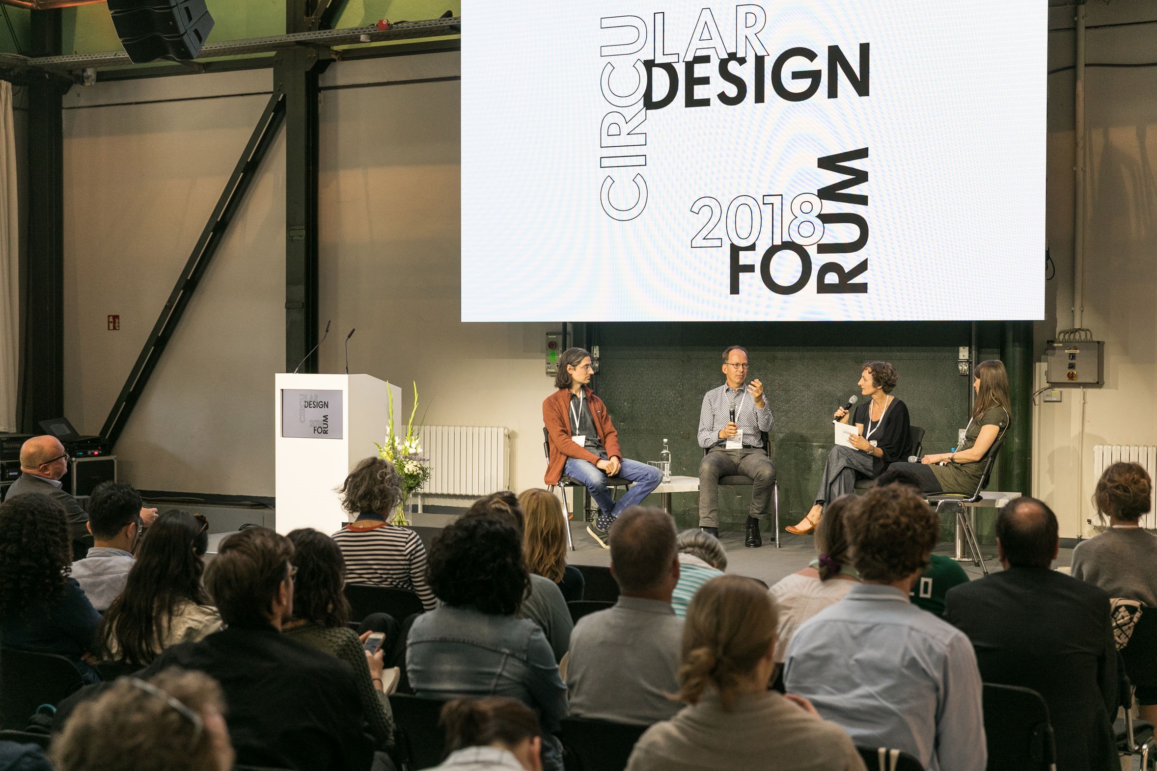 zwei Männer und zwei Frauen diskutieren mit Mikrofonen in einem Halbkreis sitzend auf einer Bühne vor Publikum, darüber ein Banner mit der Aufschrift "Circular Design Forum 2018"