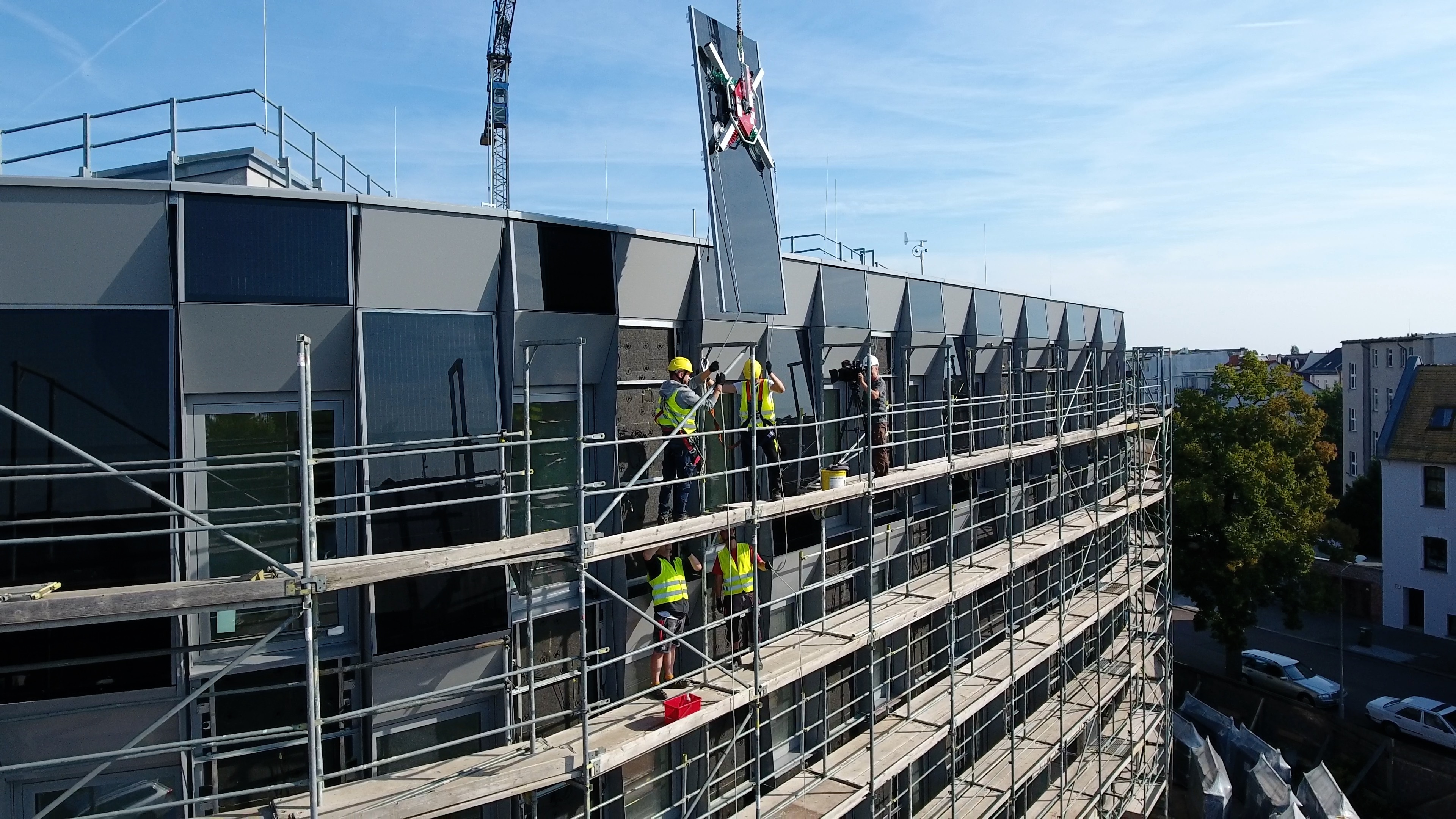 eingerüstetes Gebäude mit Fassade aus Photovoltaikmodulen, Bauarbeiter auf dem Gerüst nehmen ein Modul in Empfang, welches an einem Kran hängt