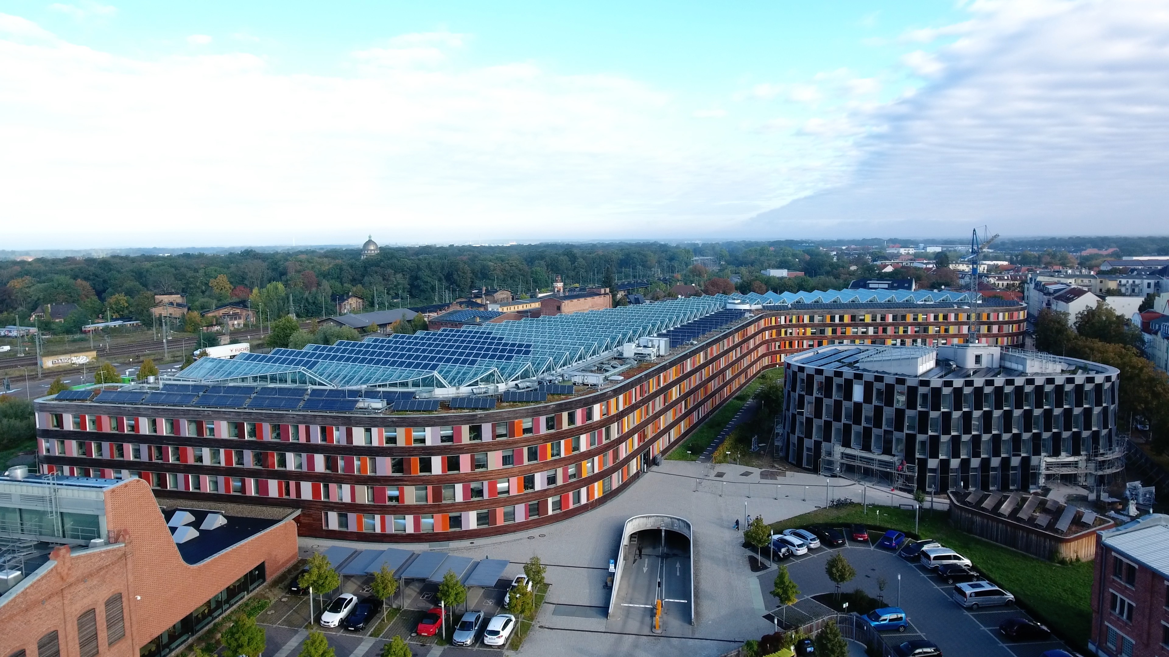 Foto von schräg oben vom Hauptgebäude des UBA Dessau mit seiner Fassade aus Holz und buntem Glas und einer Photovoltaikanlage auf dem Dach und daneben der Erweiterungbau mit seiner Fassade aus Photovoltaikmodulen