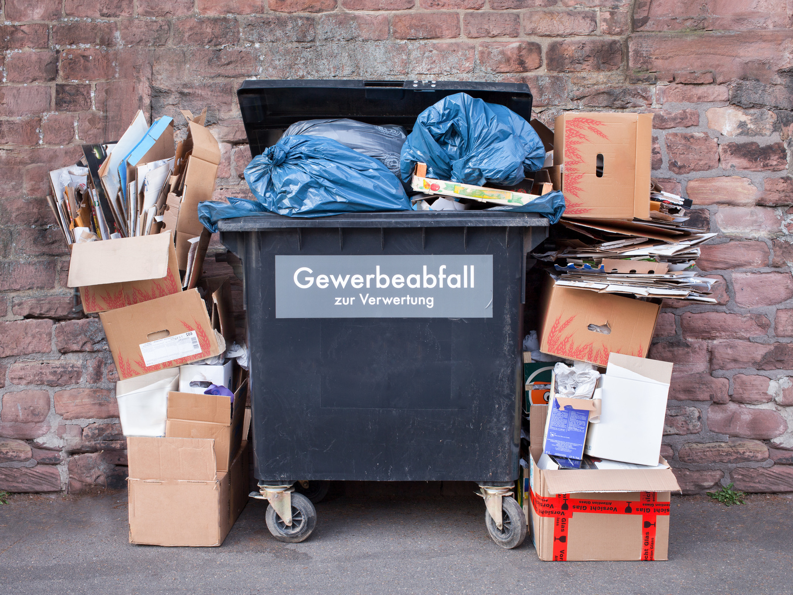 überquellender Müllcontainer in einem Hinterhof mit der Aufschrift "Gewerbeabfall zur Verwertung", daneben türmen sich leere Kartons