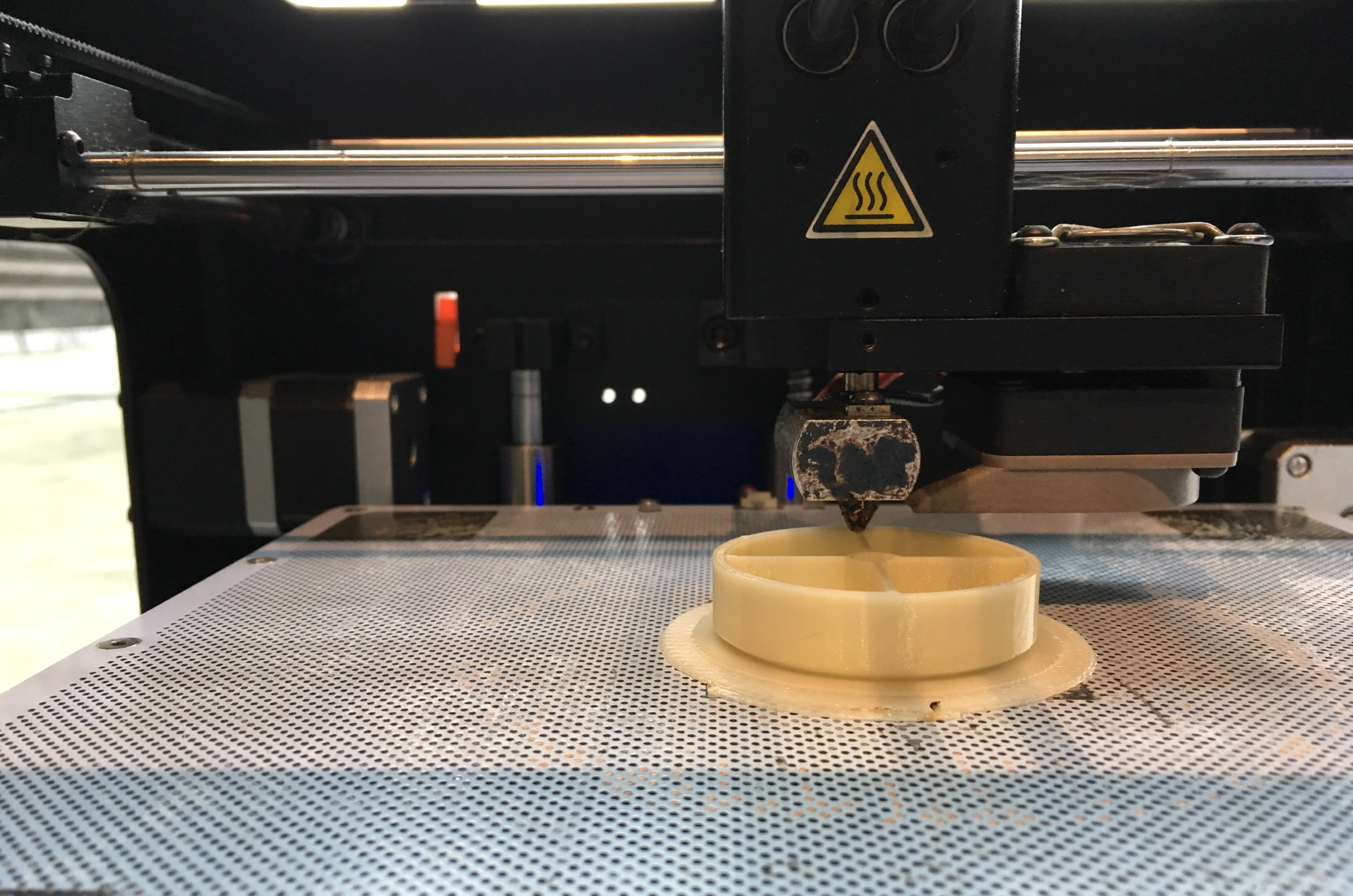 auf einer gelochten Metallplatte ist ein Plastikgegenstand zu sehen, über den der Druckkopf eines 3D-Druckers fährt