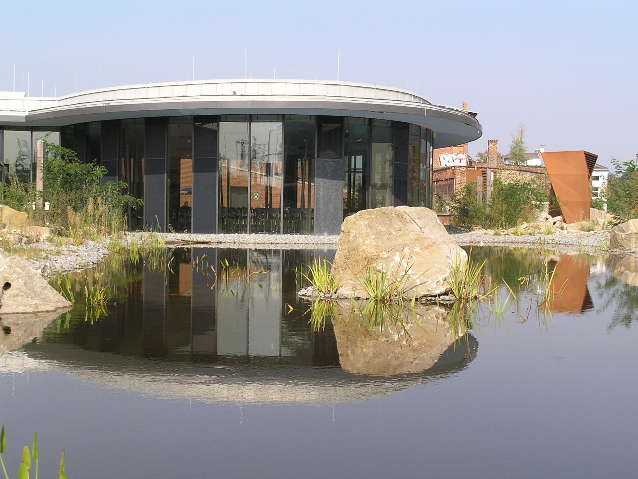 Niedriges, rundes Gebäude mit Glasfassade spiegelt sich in einem Teich mit Natursteinen