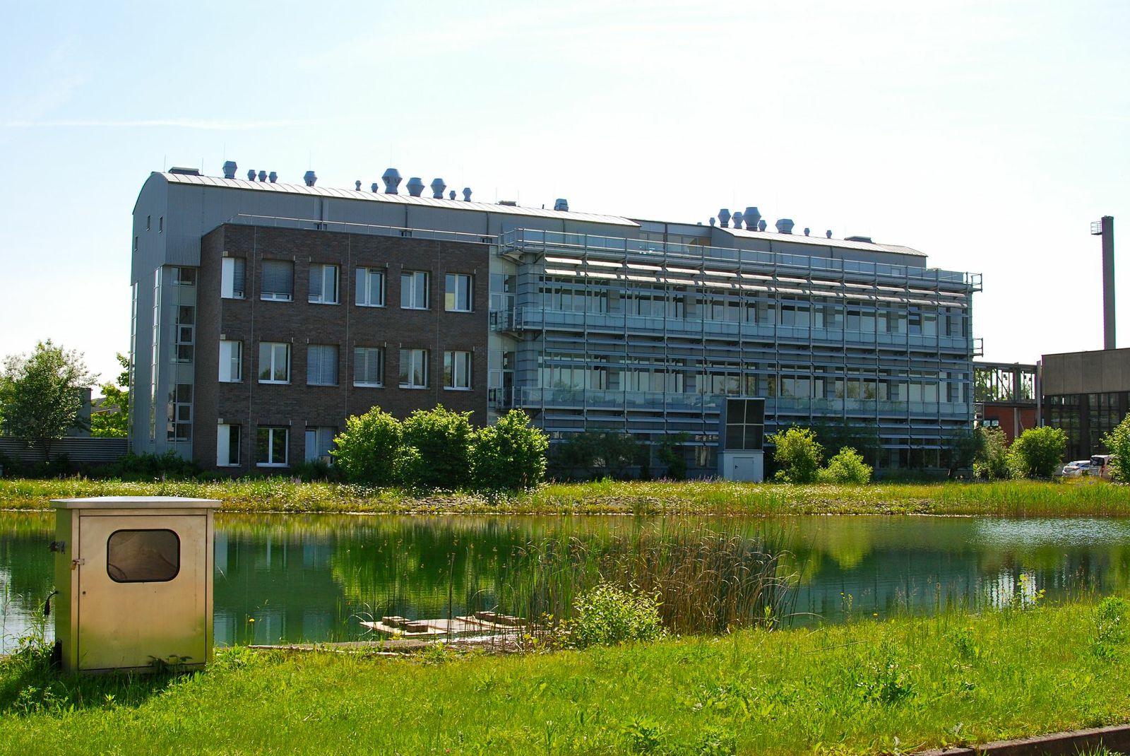 Dreistöckiges, langgezogenes modernes Bürogebäude, davor ein Fluss oder Kanal mit grasbewachsenen Ufern