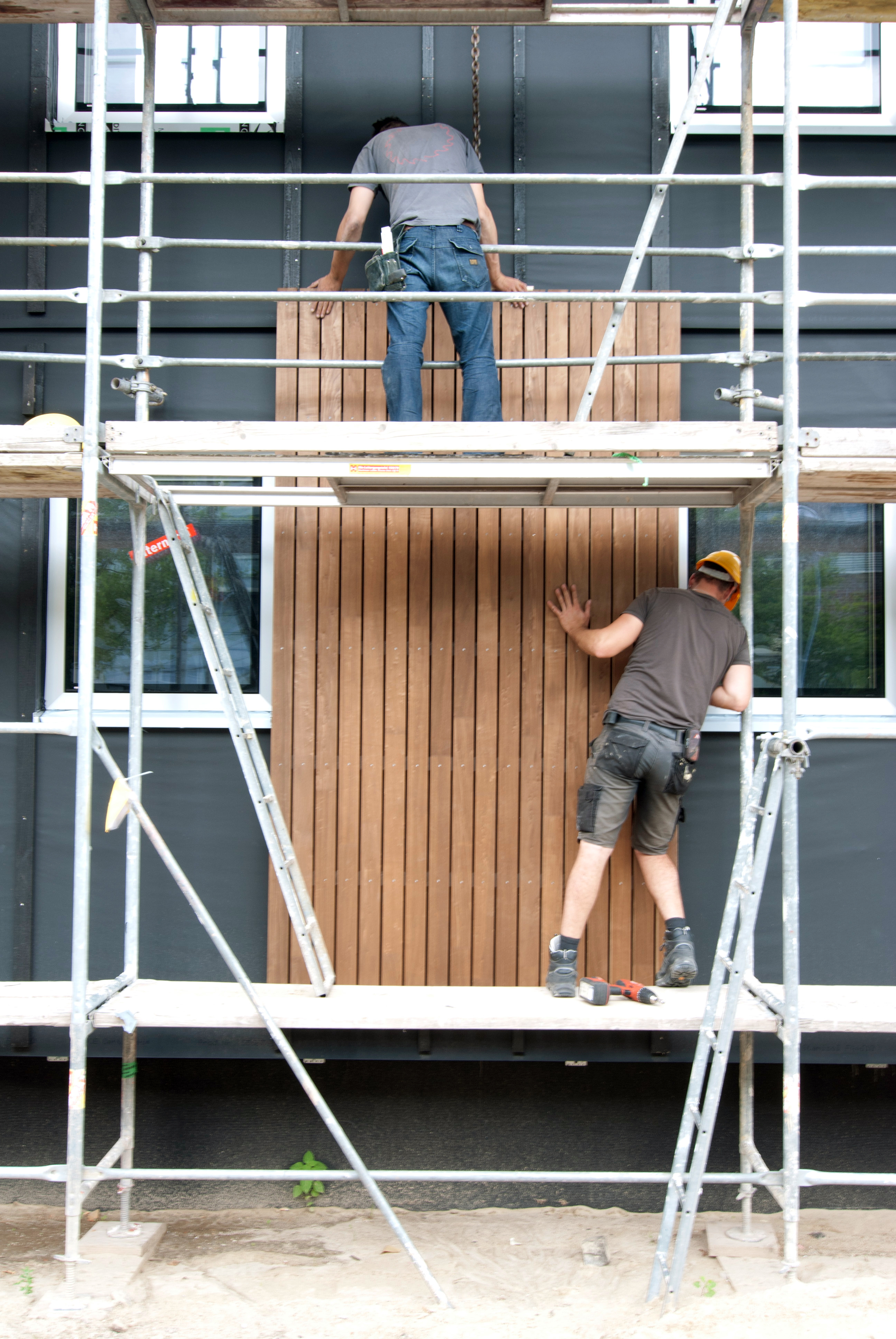 zwei Bauarbeiter stehen auf einem Baugerüst und montieren zusammen ein Holz-Fassadenelement
