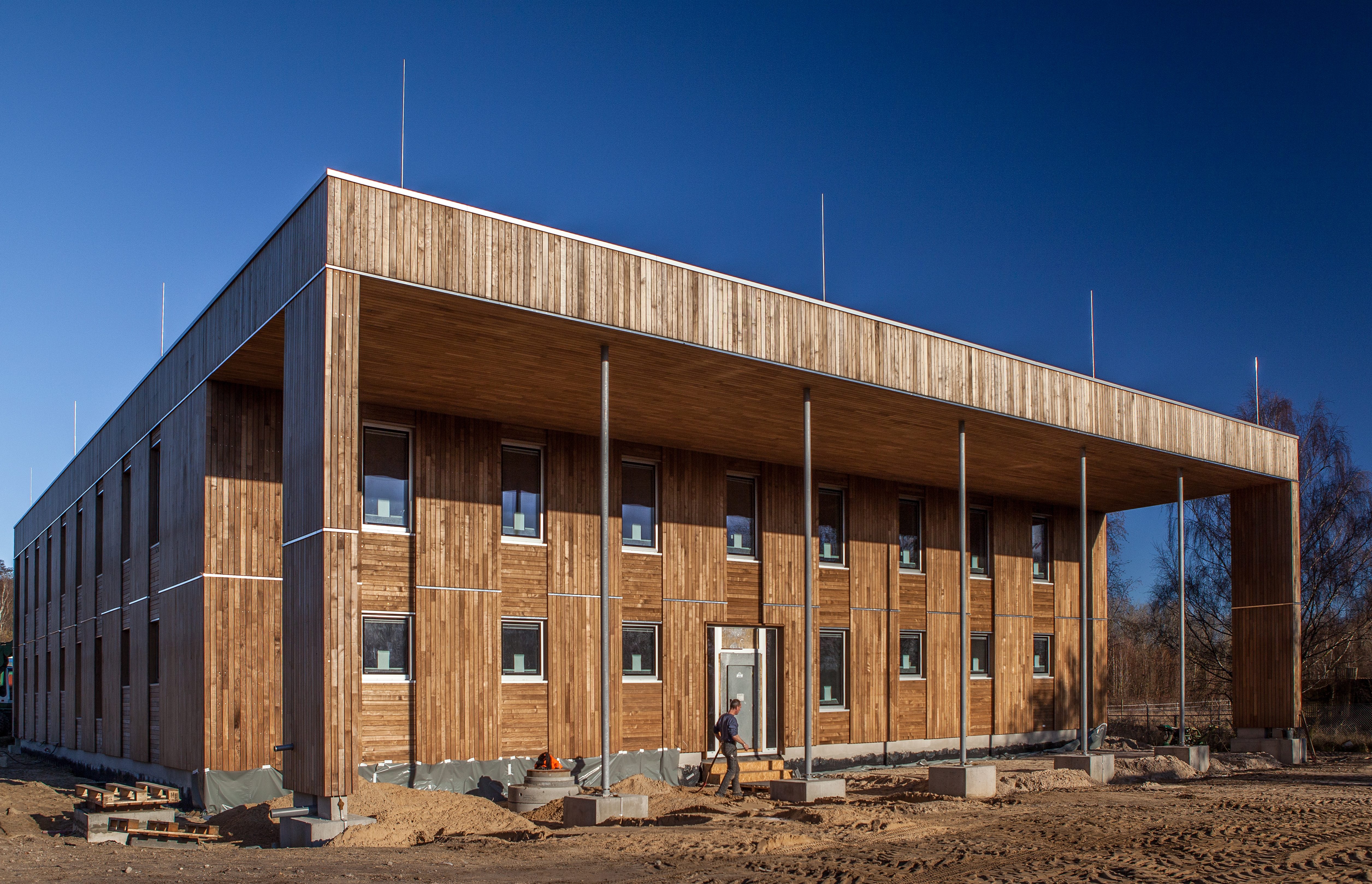 würfelförmiger, zweistöckiger Bau mit Holzfassade und Kollonadengang, davor ein Bauarbeiter, die Außenanlage noch ungestaltete Baustelle