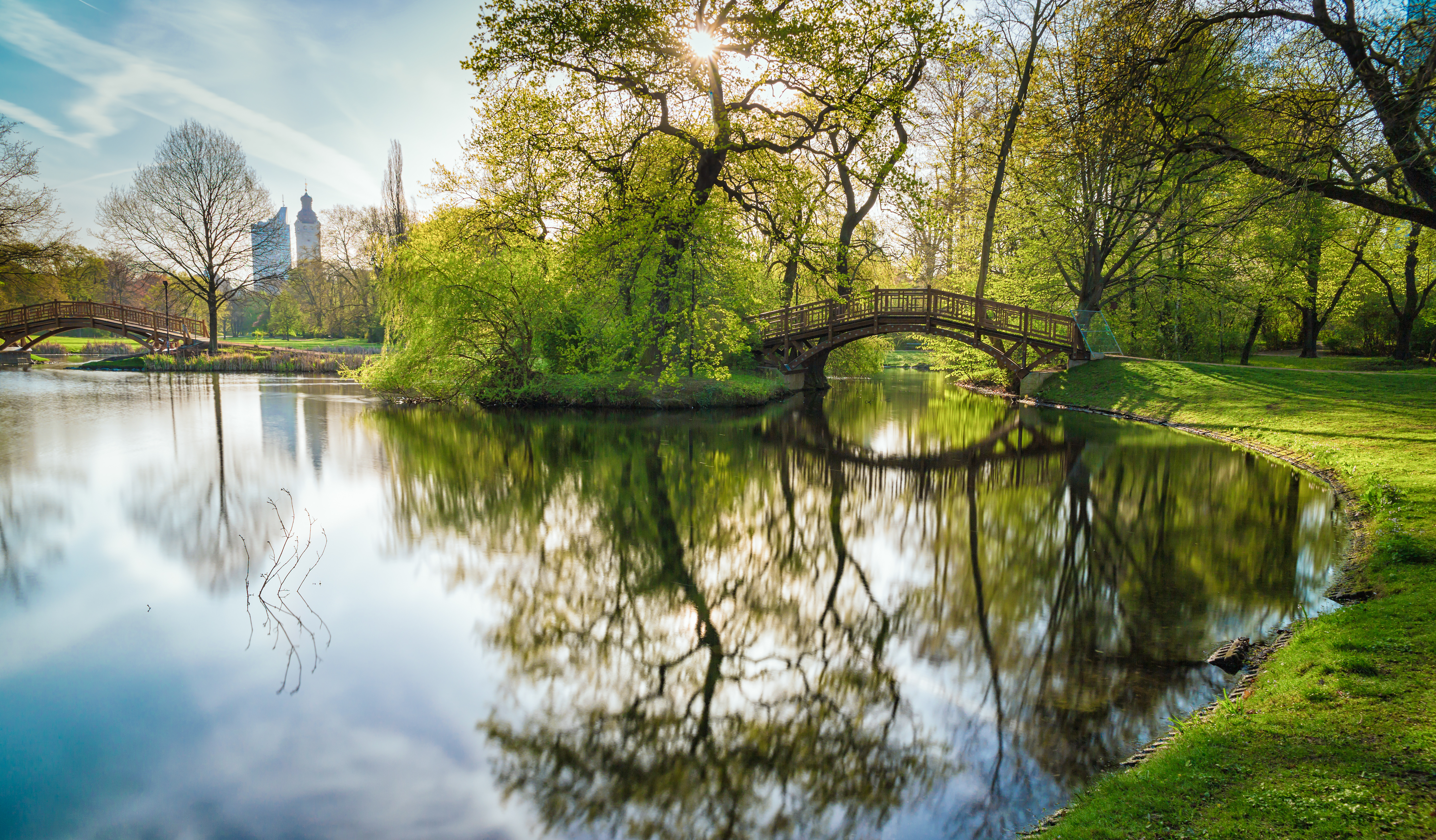 in einem Park spiegeln sich Bäume und eine Brücke in einem Teich, im Hintergrund Gebäude einer Großstadt
