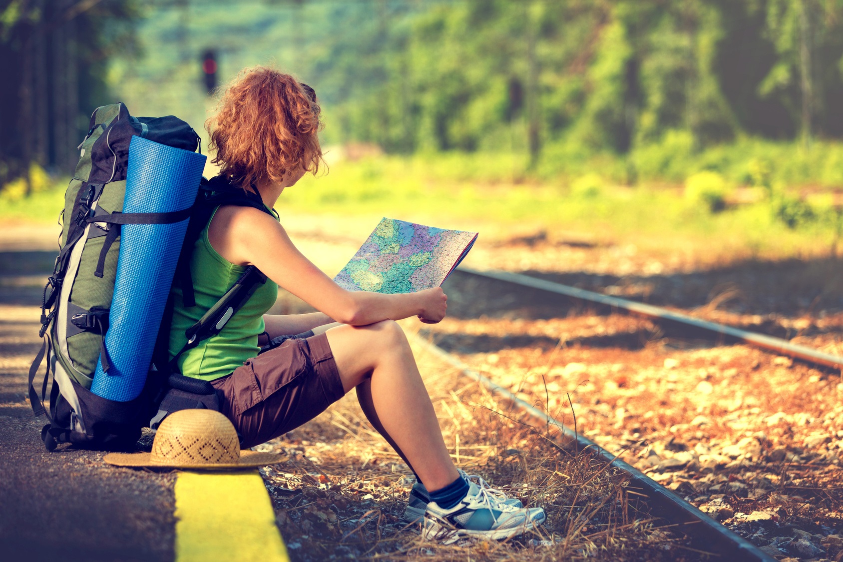 eine junge, sportlich gekleidete Frau mit Rucksack, Strohhut und Isomatte wartet auf der Bahnsteigkante in der Sonne sitzend auf einen Zug und schaut in eine Landkarte