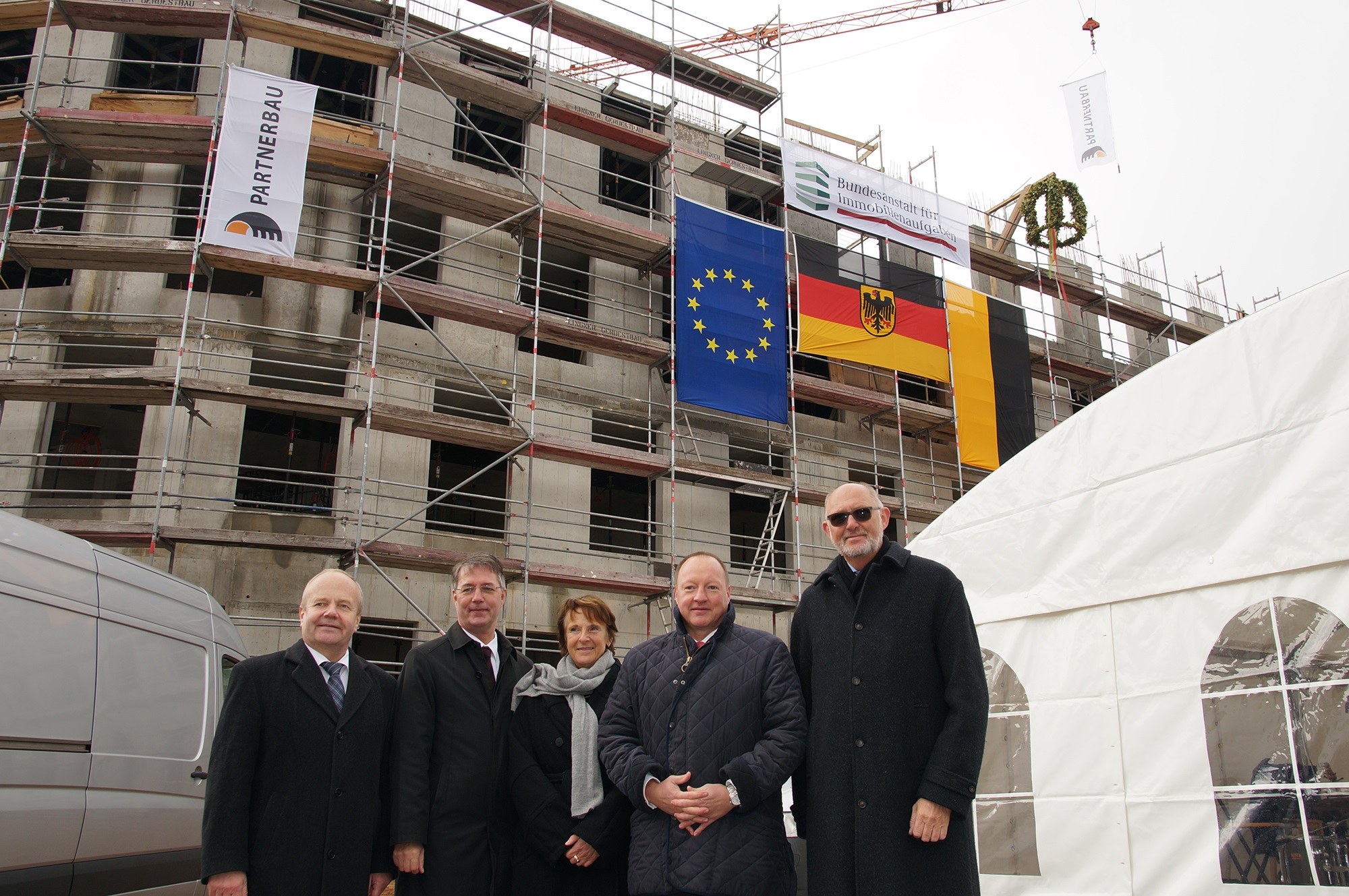 Gruppenfoto: vier Männer und eine Frau vor einem eingerüsteten Rohbau mit Richtkranz und Fahnen der EU, Deutschland, Sachsen-Anhalt, Bundesanstalt für Immobilienaufgaben und Partnerbau