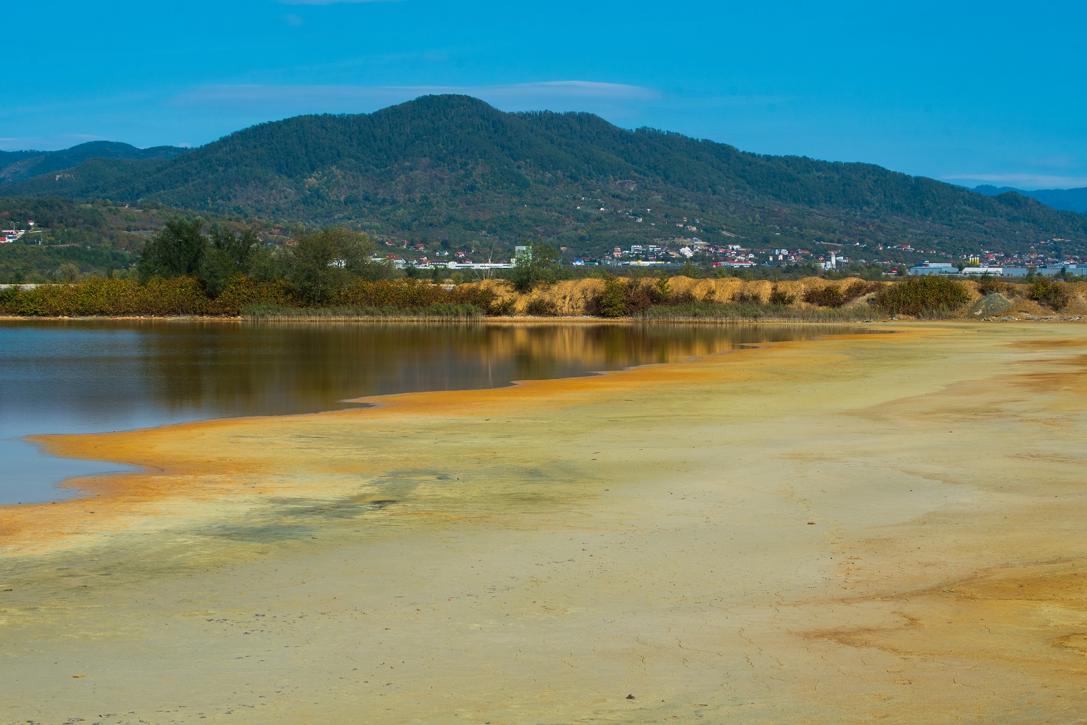 zum Teil wassergefülltes Rückhaltebecken mit naturnahem Ufer, im Hintergrund bewaldete Berge