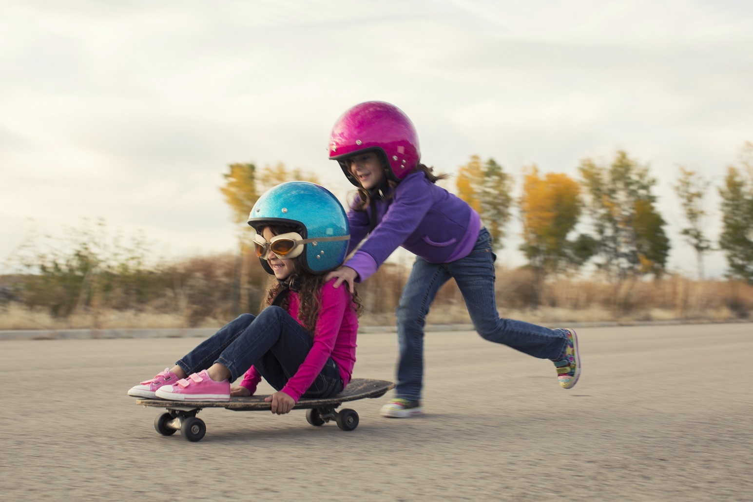 zwei kleine Mädchen mit Sturzhelmen schieben sich gegenseitig auf einem Skateboard auf einem gepflasterten Platz