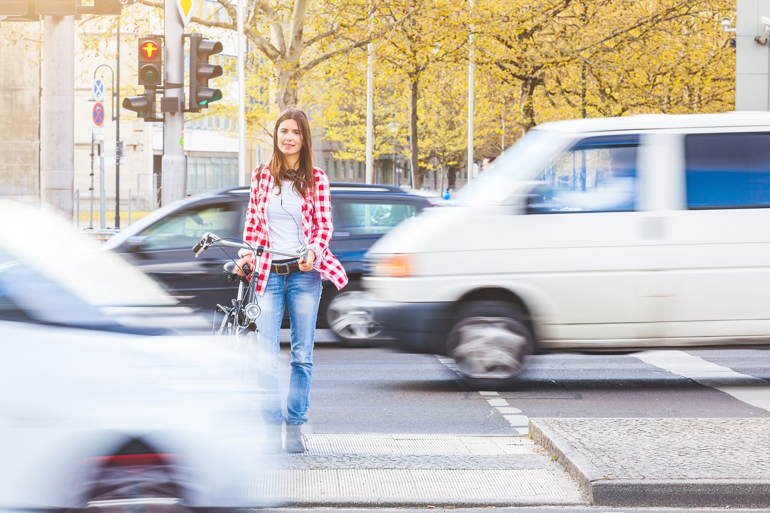 eine junge Frau wartet, ein Fahrrad schiebend, auf einer Mittelinsel an einer roten Ampel, vor und hinter ihr fahren Autos vorbei