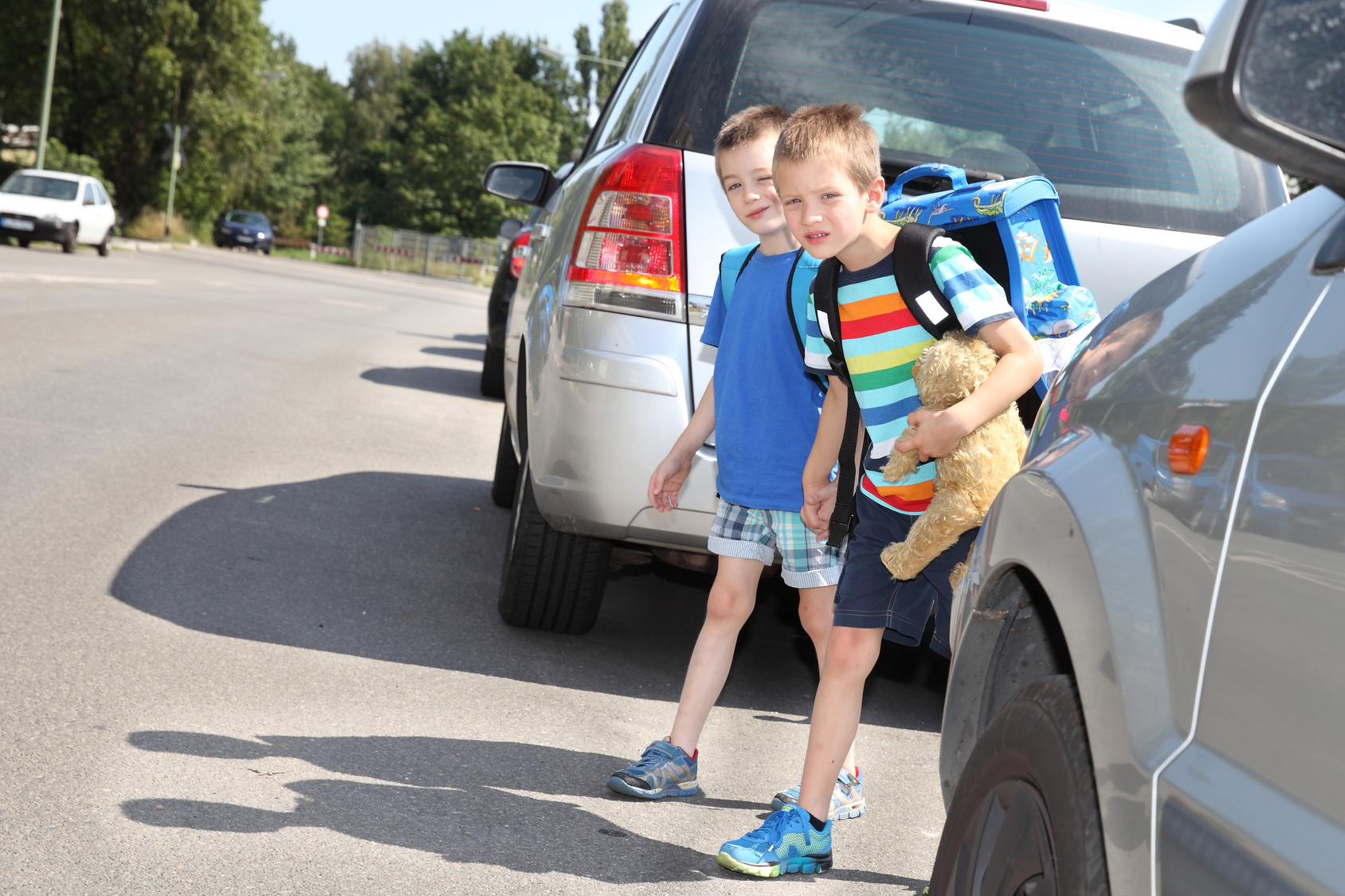 zwei Kinder mit Schulranzen schauen zwischen parkenden Autos, ob sie die Straße sicher queren können