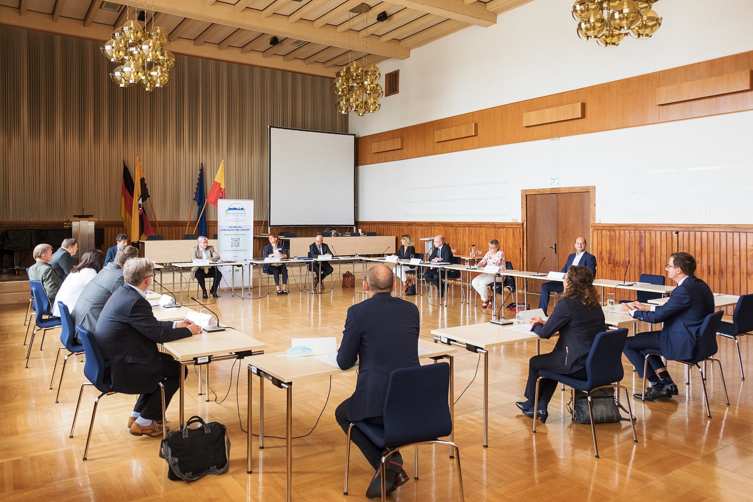 in einem Saal sitzen Männer und Frauen an Tischen im Kreis, hinten ein Aufsteller und Fahnen