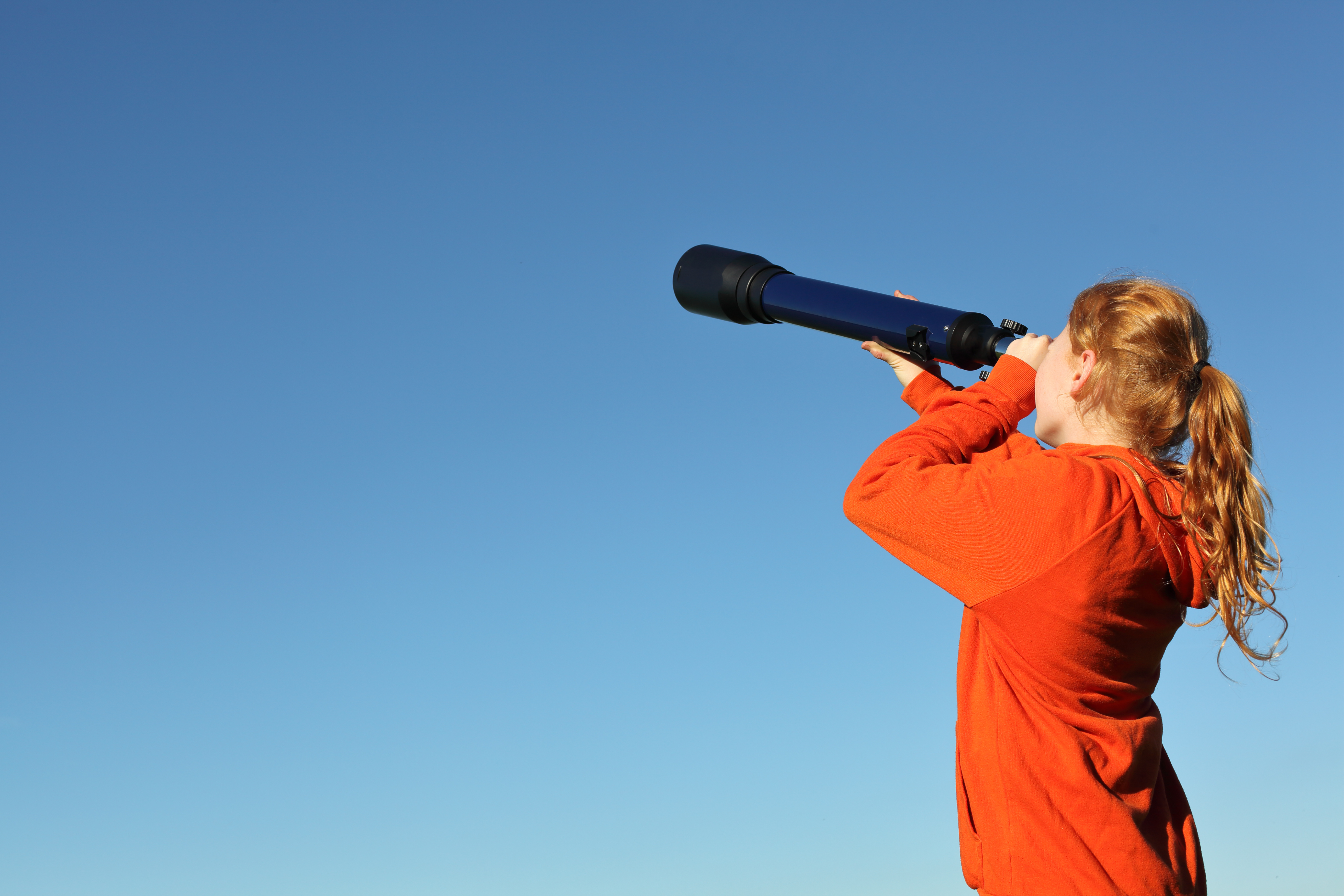 eine junge Frau schaut mit einem Fernrohr in die Ferne, im Hintergrund klarer, blauer Himmel