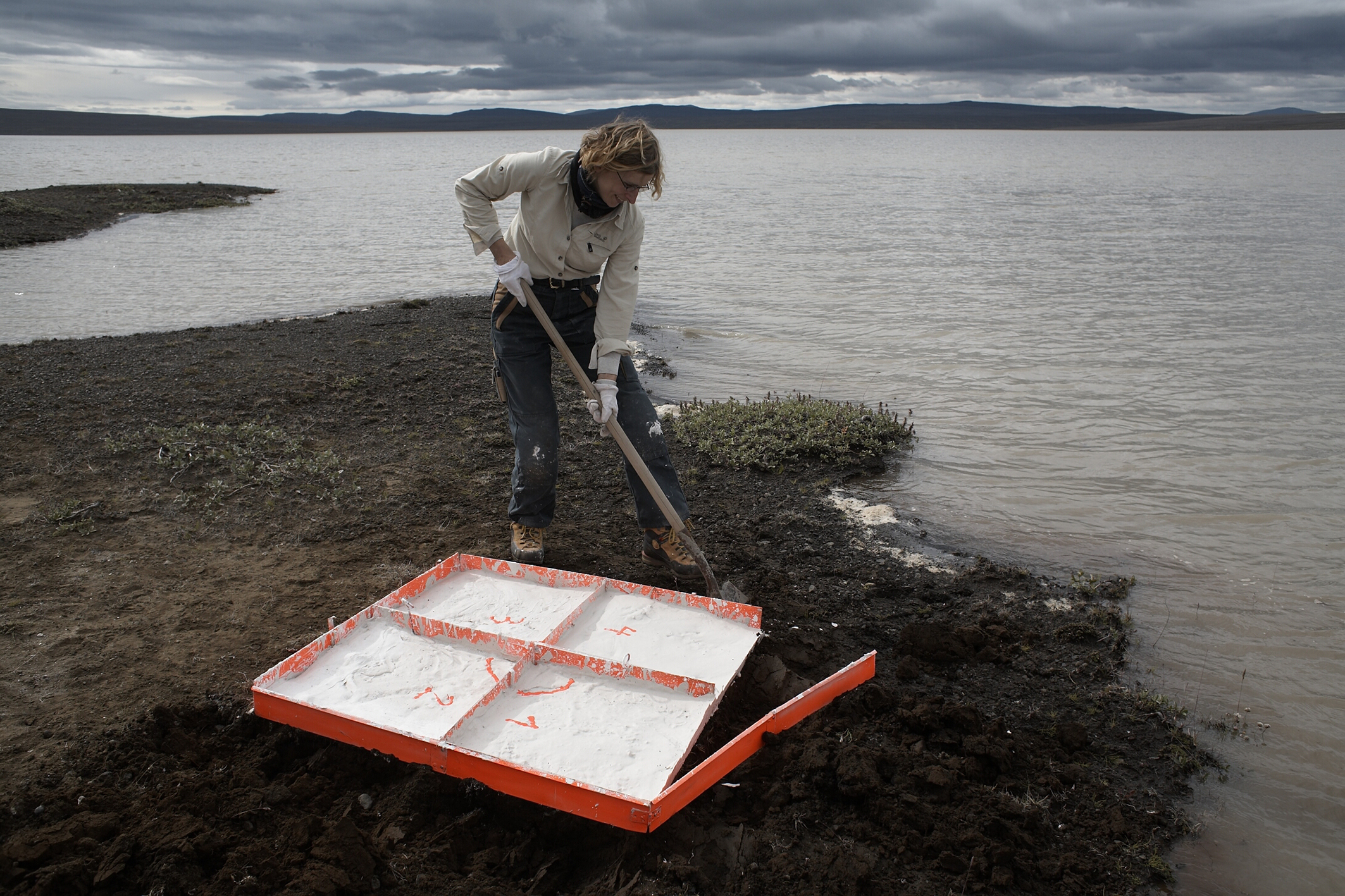 Die Künstlerin steht an Ufer eines Sees und nimmt eine Bodenprobe. 