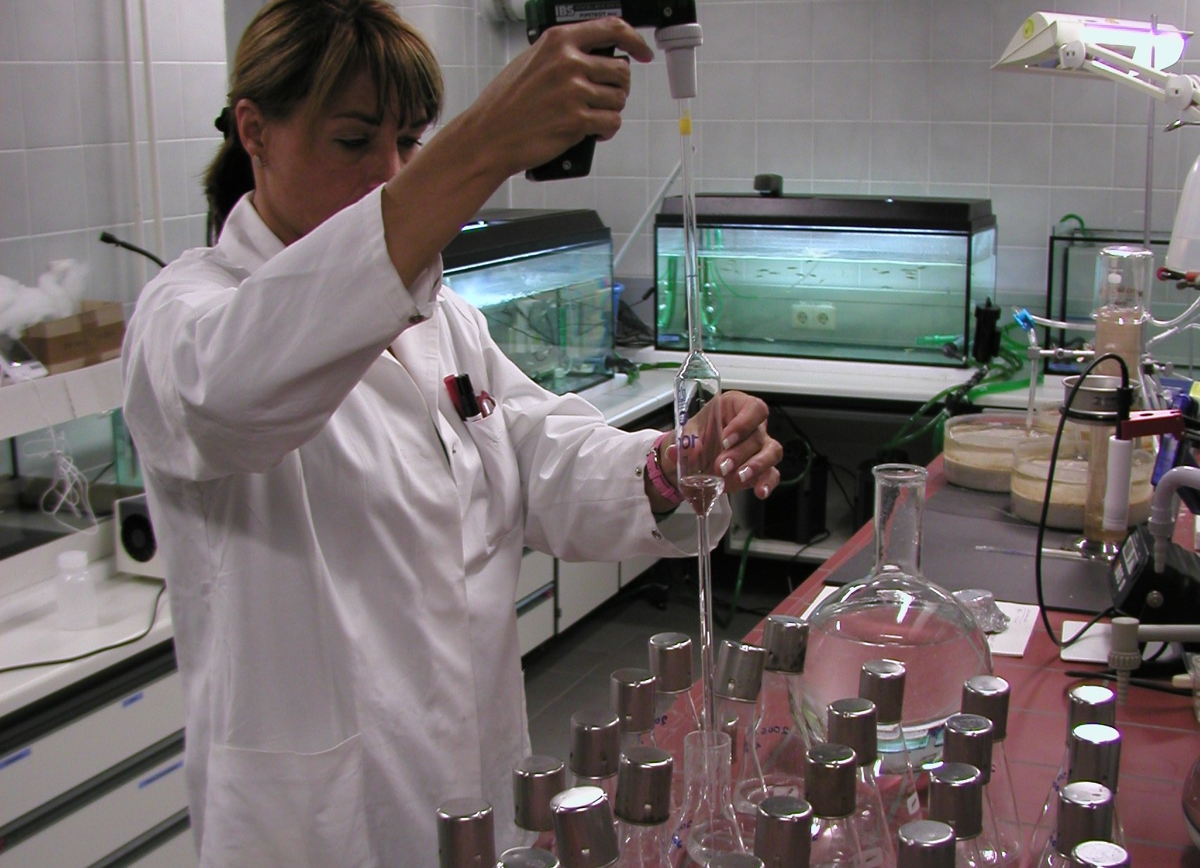Eine Frau im Chemiekittel arbeitet im Labor. Sie befüllt Glasgefäße mit Flüssigkeit.