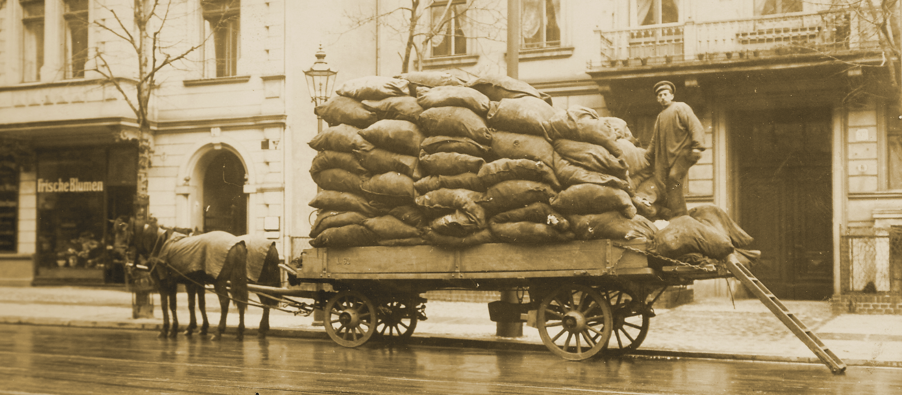 Ein Arbeiter steht auf der Ladefläche eines Pferdewagens, auf der viele Säcke gestapelt sind. Davor steht eine Leiter, über die er auf den Wagen gekommen ist. 