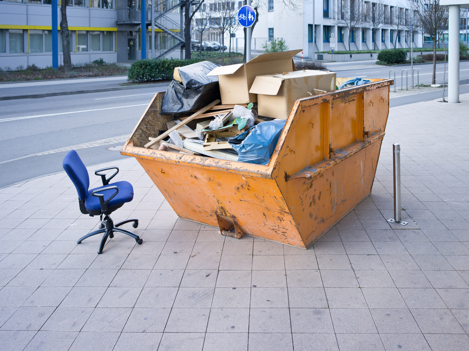 Ein Müllcontainer steht auf der Straße, daneben steht ein Bürostuhl