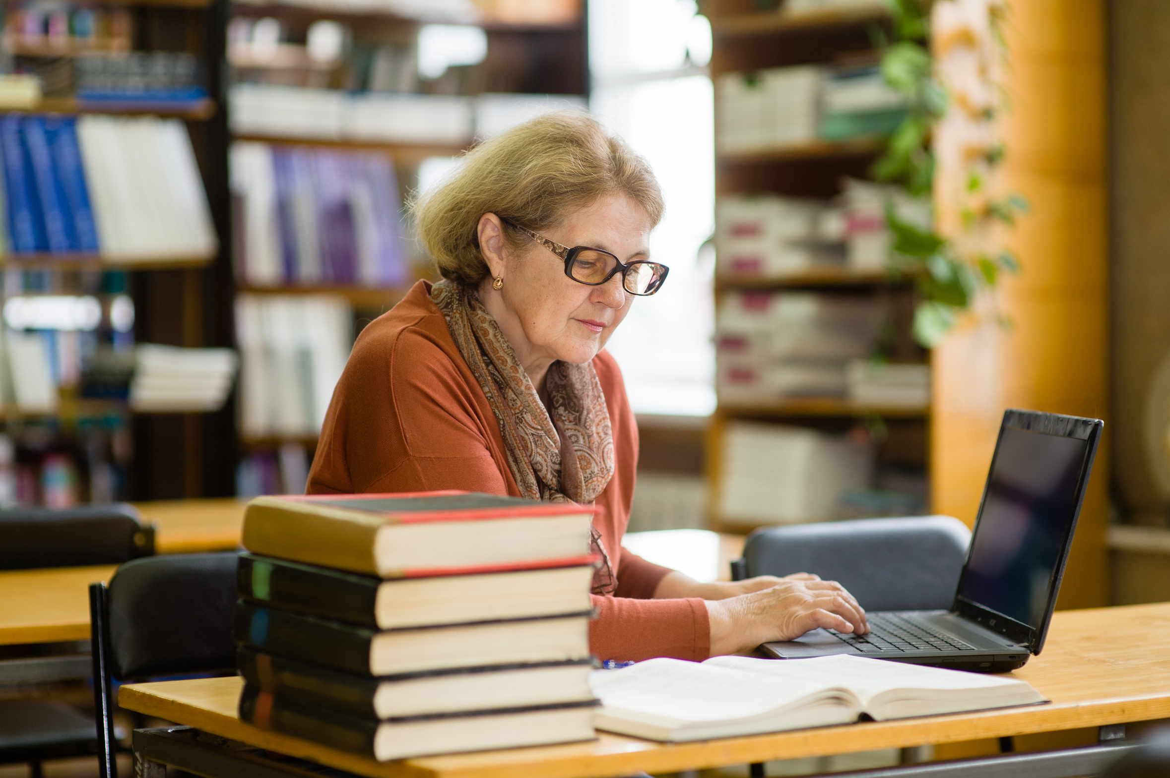 Eine Frau sitzt mit einem Laptop in einer Bibliothek und liest ein Buch.