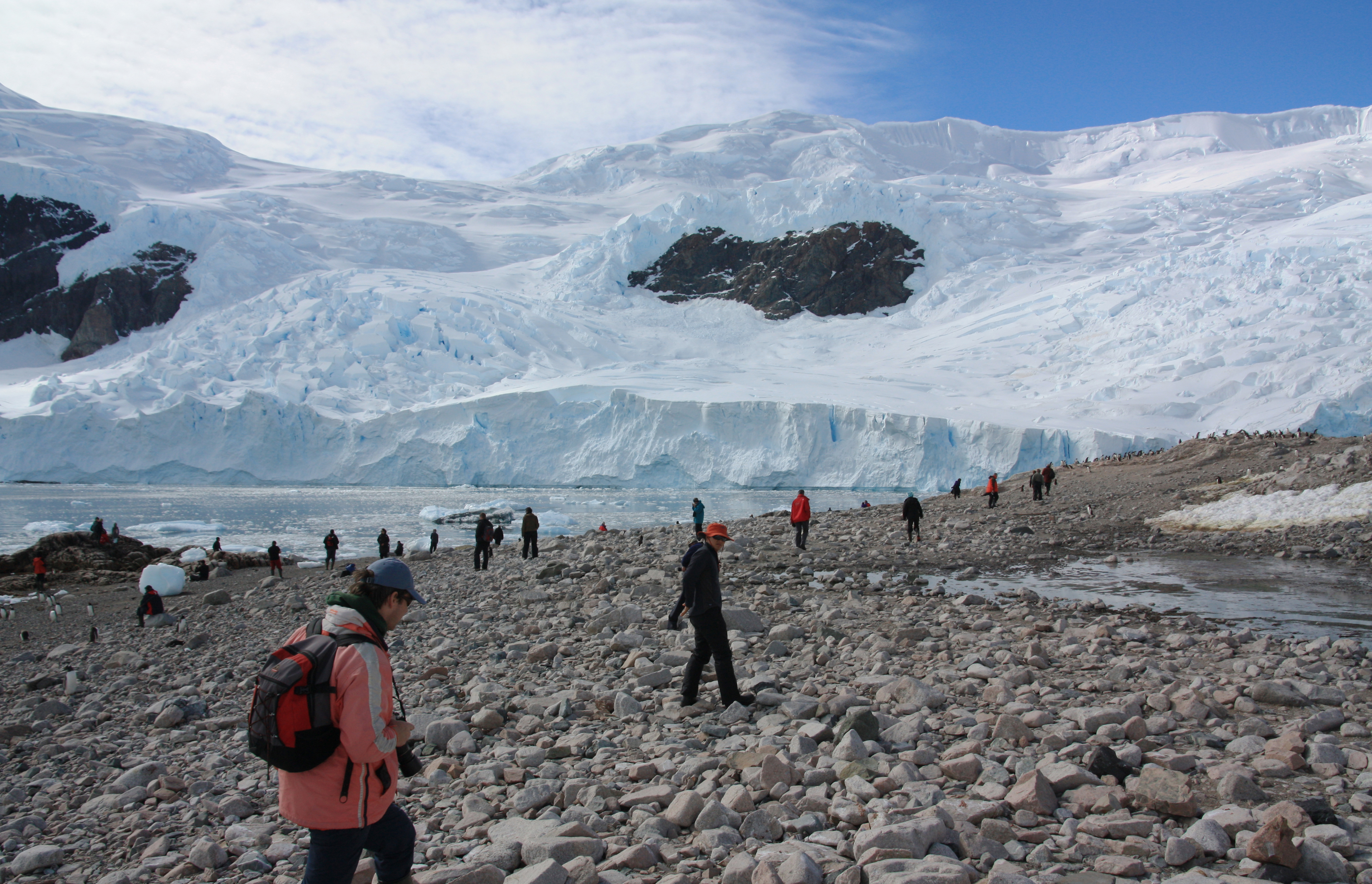 Im Vordergrund laufen Touristen in Funktionskleidung auf steinigem Strand. Im Hintergrund sieht man große Eismassen.