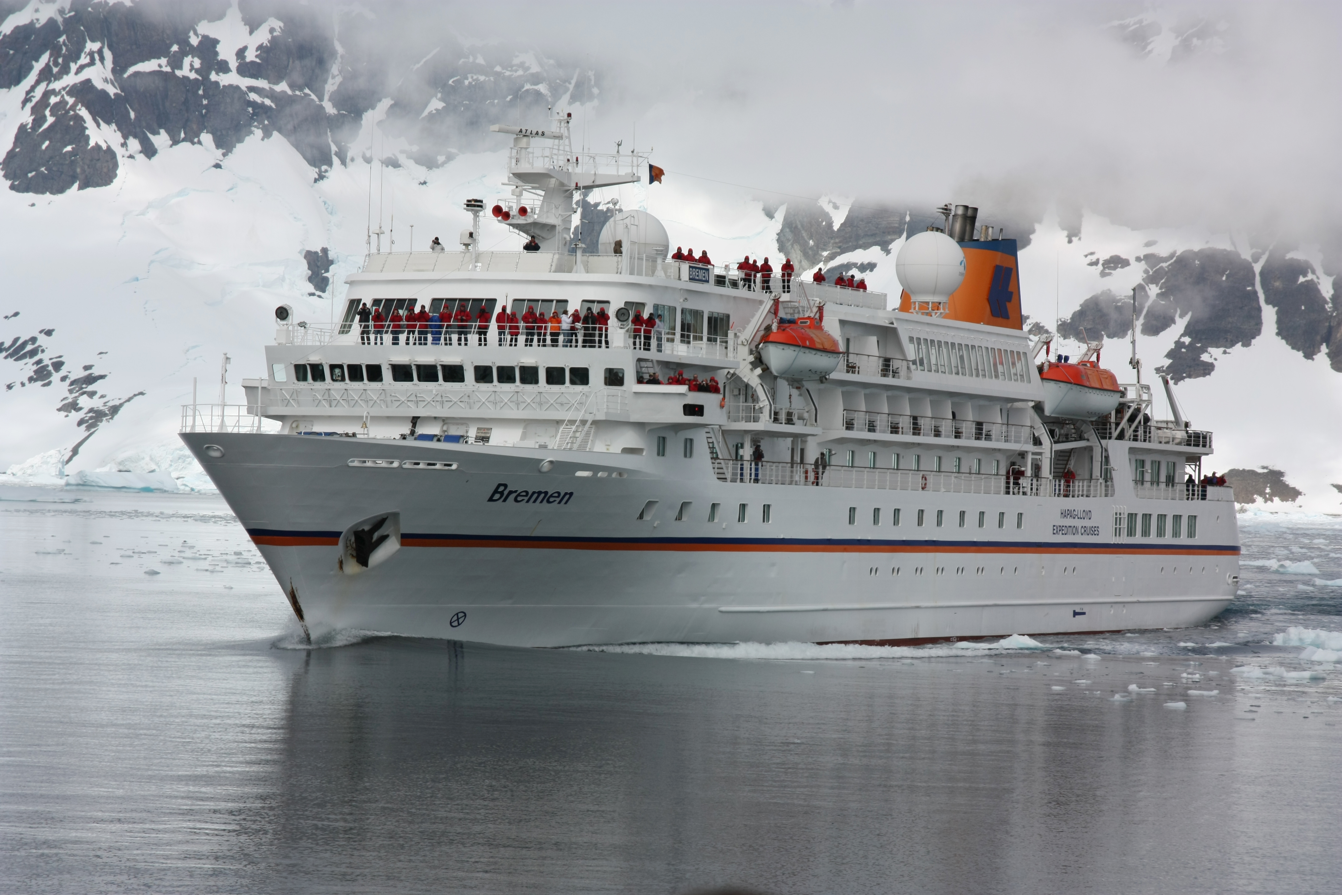 Das Kreuzfahrtschiff Bremen fährt durch antarktisches Gewässer. Hinter ihr ragt eine steile schneebedeckte Felswand auf. Die Wolken hängen sehr tief. Auf dem Schiff stehen Menschen in Kälteschutzanzügen. 