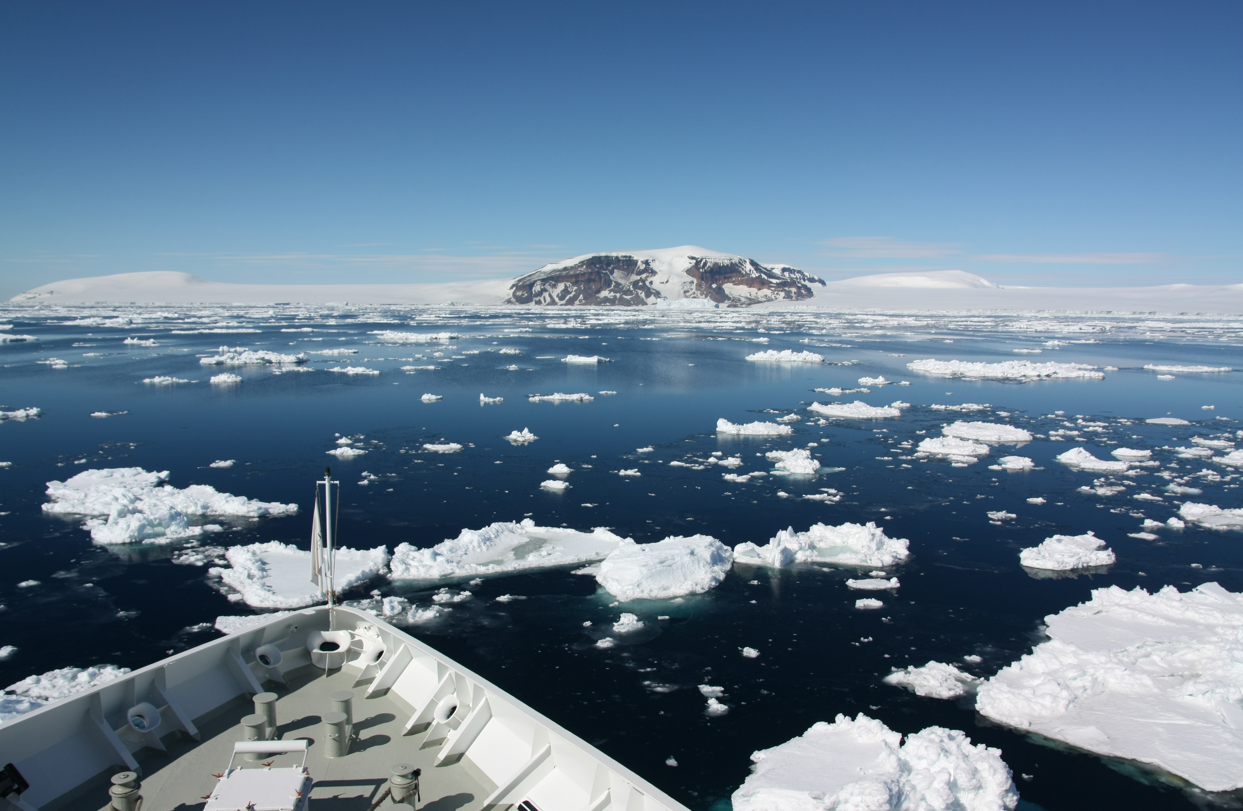 Die Aufnahme ist vom Bug eines Schiffes gemacht, so sind noch der vorderste Teil des Schiffes und dann das Südpolarmeer im Bild. Auf dem Wasser treiben Eisschollen und im Hintergrund ist antarktisches Festland. 