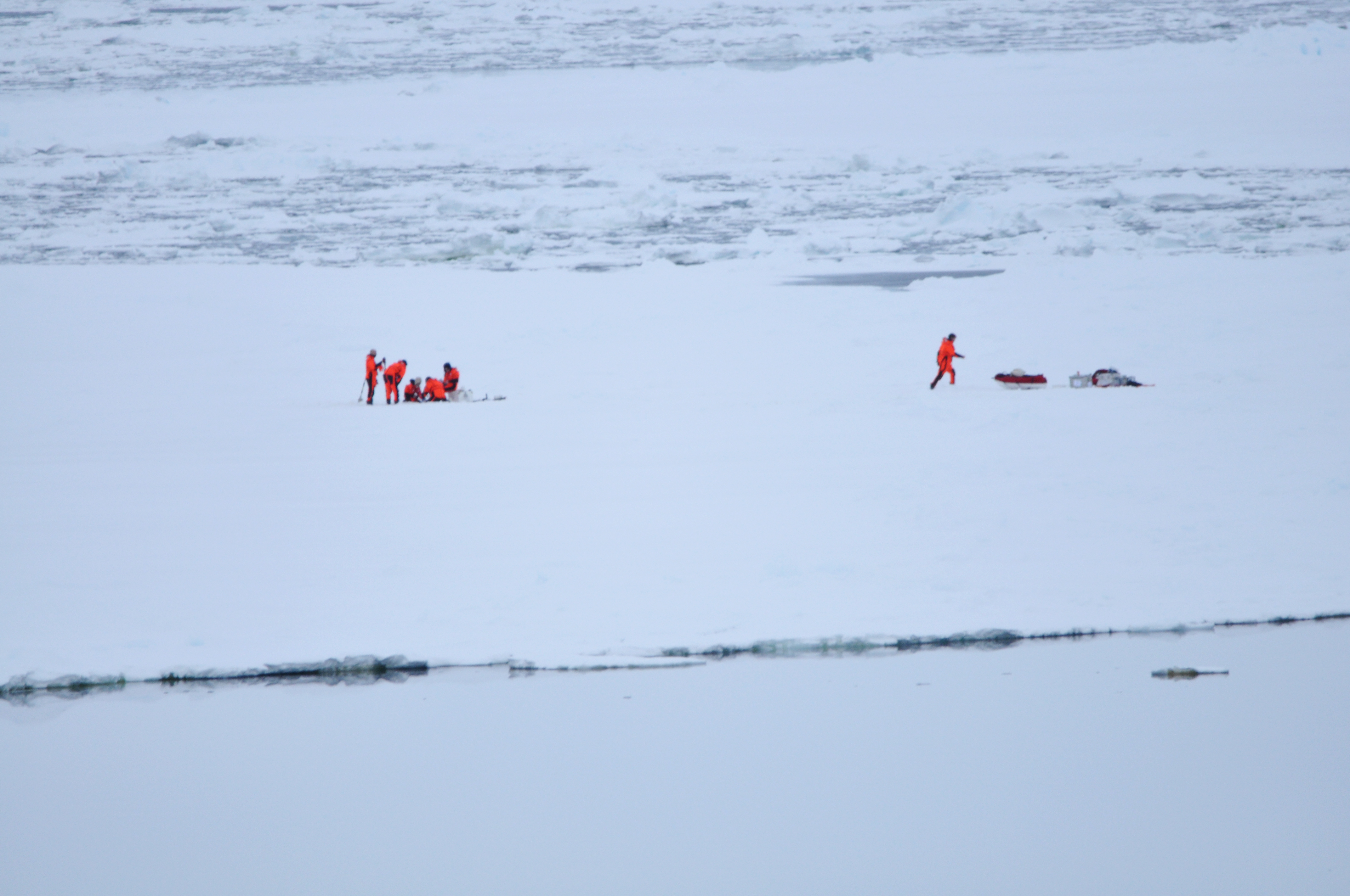 Auf einer weiten Eisebene steht eine Gruppe von Forschern und roten Kälteschutzanzügen. Sie entnehmen an einer Stelle eine Eisprobe. Weiter rechts läuft eine weitere Person zur Ausrüstung. 